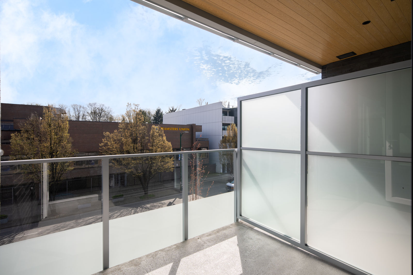 A modern balcony with glass railings and frosted privacy panels overlooks a street with trees and commercial buildings on a clear day.