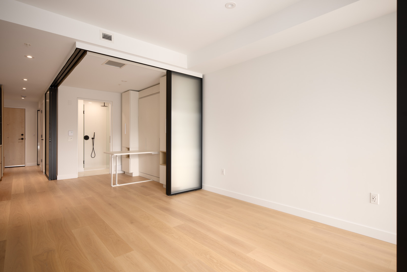 Empty modern room with light wood flooring, white walls, and a small open area with a folding table and sliding glass partitions near an accessible shower and entryway.
