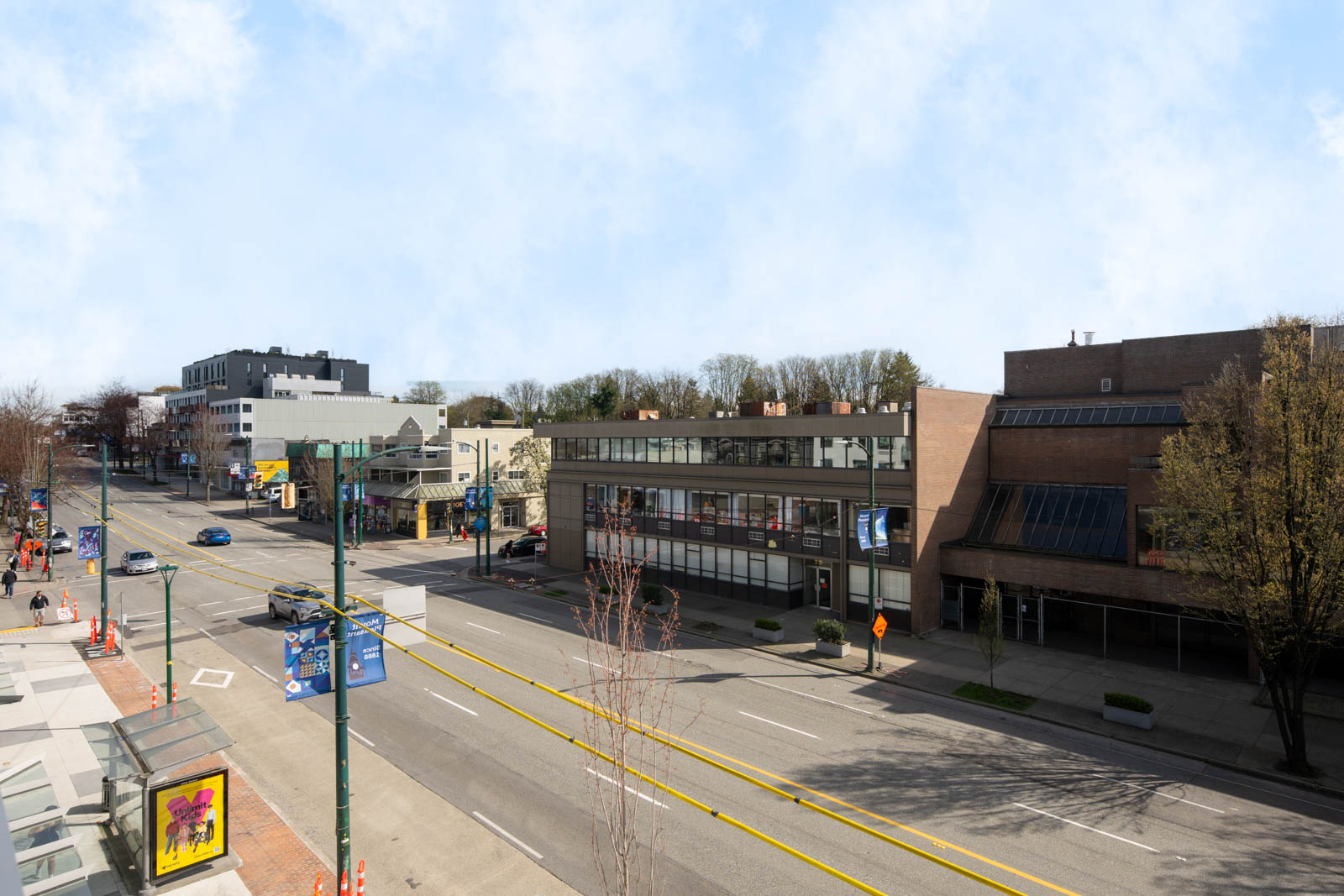 A quiet city street with low-rise buildings, trees, parked cars, and banners on light poles under a partly cloudy sky.