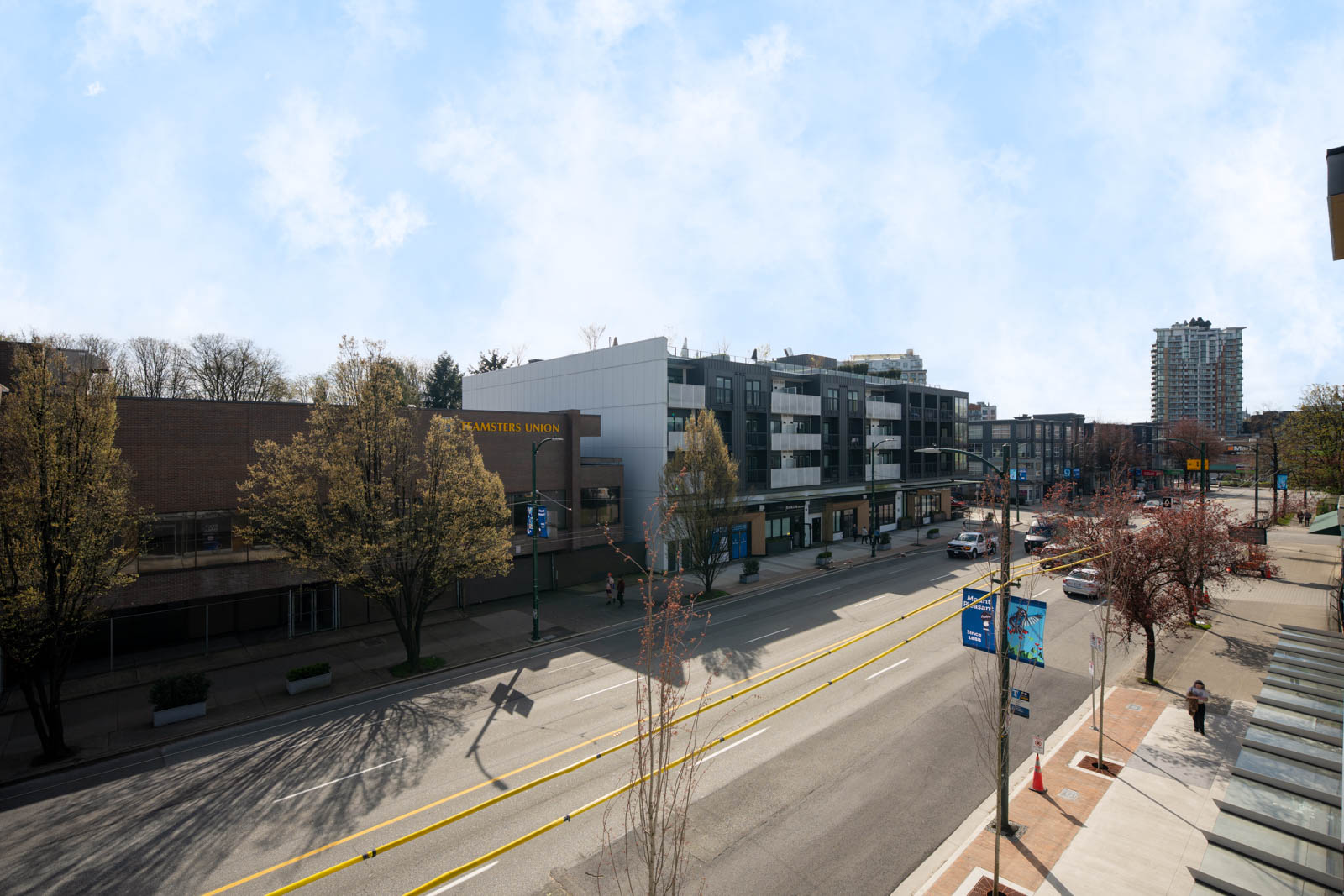 A wide street lined with trees and modern buildings on a clear day, with minimal traffic and a few pedestrians on the sidewalk.