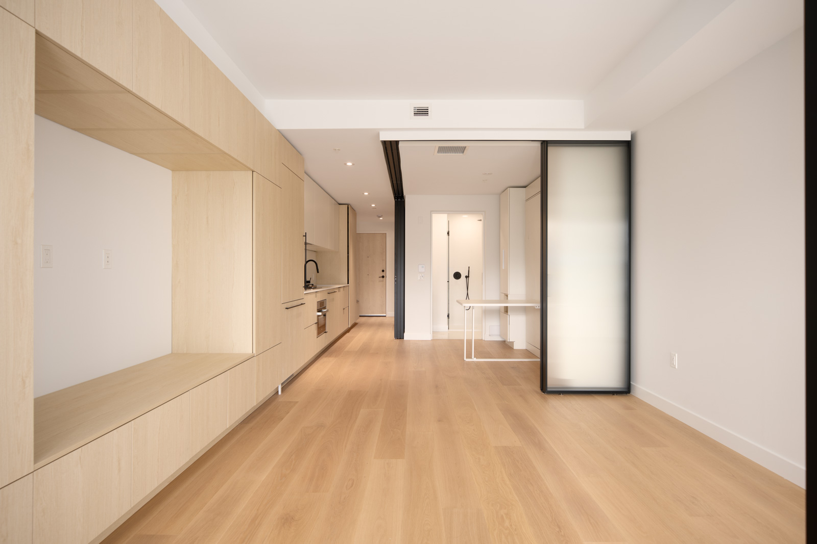 Modern, unfurnished apartment with light wood floors and cabinetry, built-in shelving, white walls, and a small kitchen area. A glass divider separates the space near the entrance.
