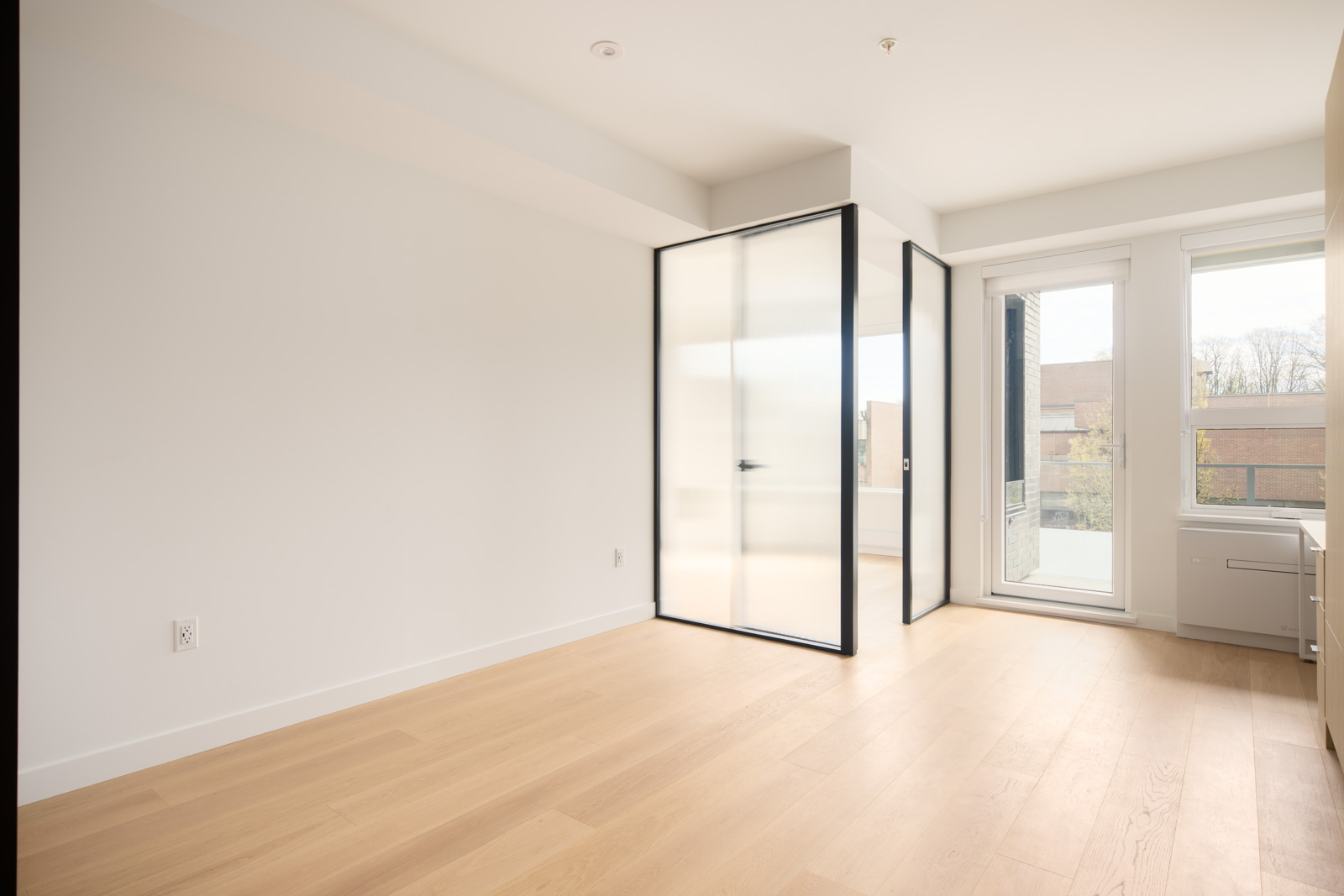 An empty, modern room with light wood floors, white walls, glass partition doors, and large windows allowing natural light inside.