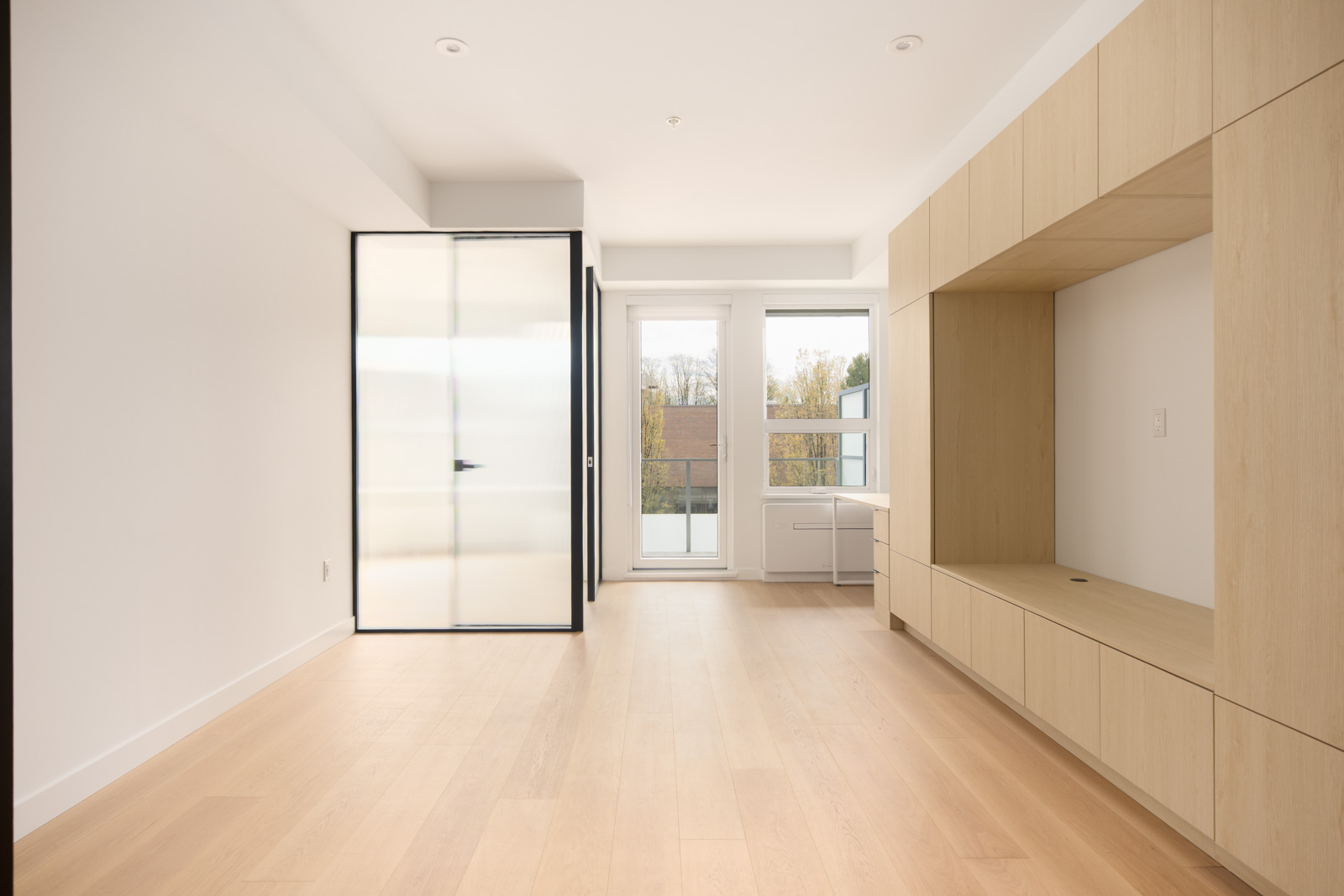 Minimalist empty room with light wood floors, built-in wooden cabinets, frosted glass door, and large windows letting in natural light.