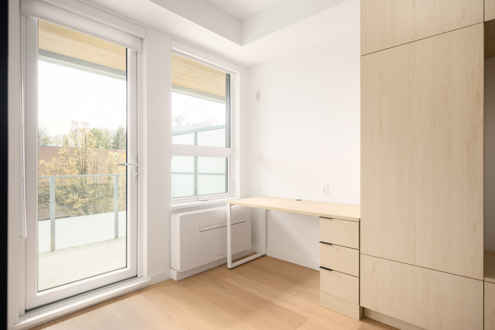Minimalist room with light wood flooring, built-in desk and drawers, large windows, a glass door to a balcony, and neutral walls.