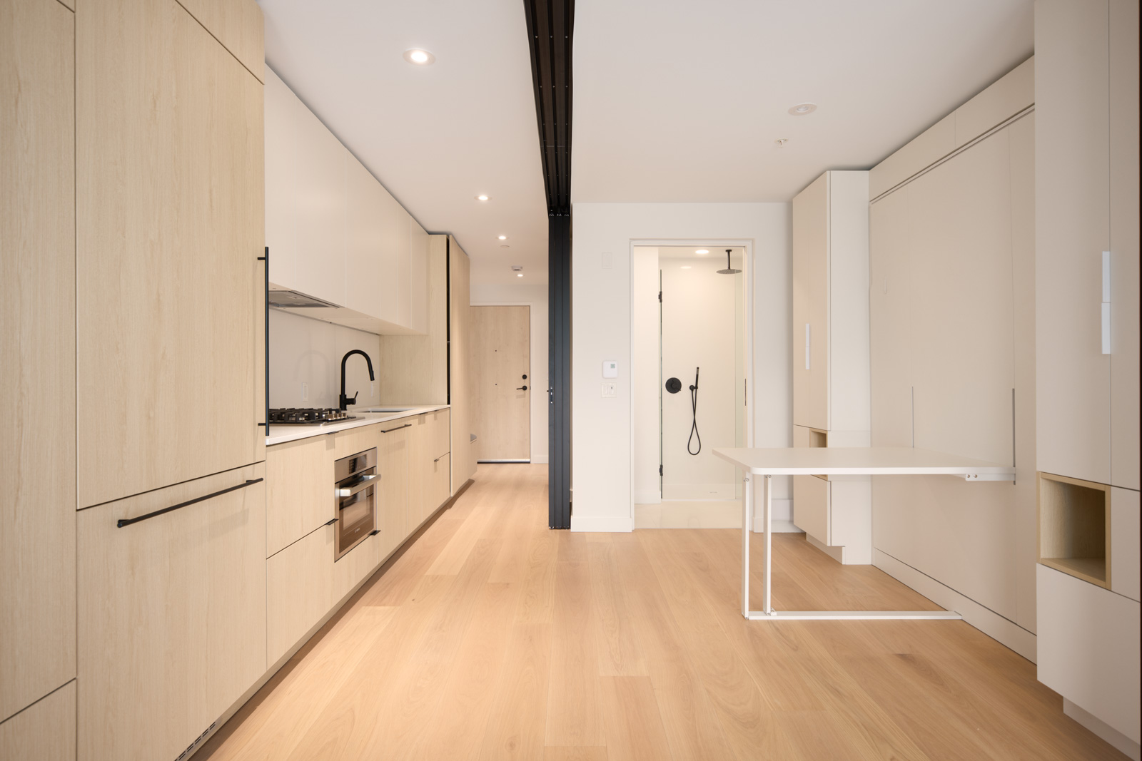 Modern minimalist kitchen with light wood cabinets, built-in appliances, light wood flooring, a fold-down table, and a compact bathroom visible in the background.