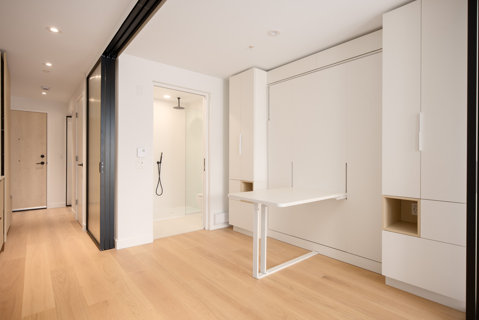 Minimalist apartment interior with light wood flooring, white walls, built-in storage cabinets, a fold-down table, and a bathroom with a glass shower door visible in the background.