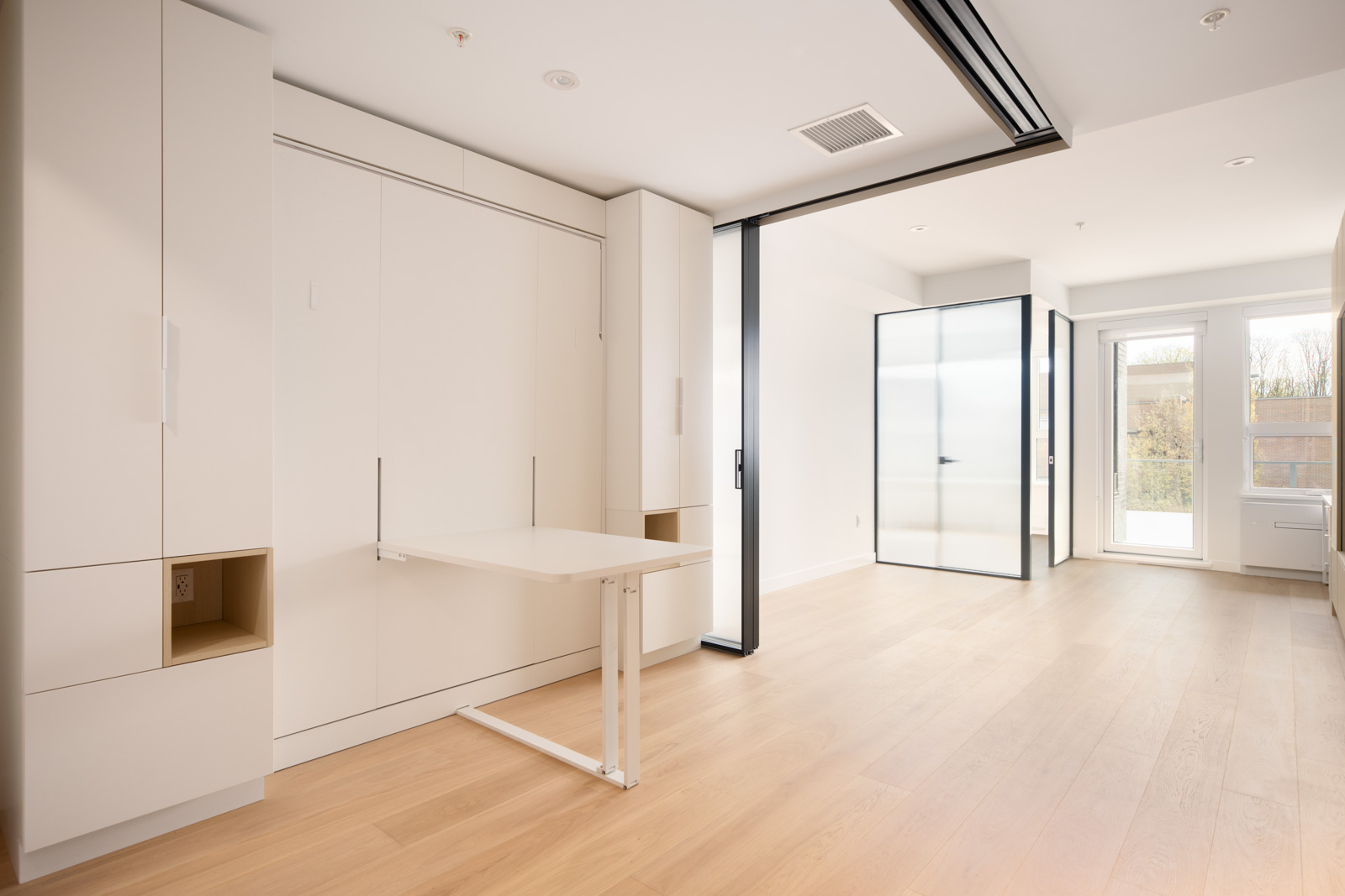 A modern, minimalist room with light wood floors, built-in white cabinetry, a fold-out table, large windows, and glass partition walls.