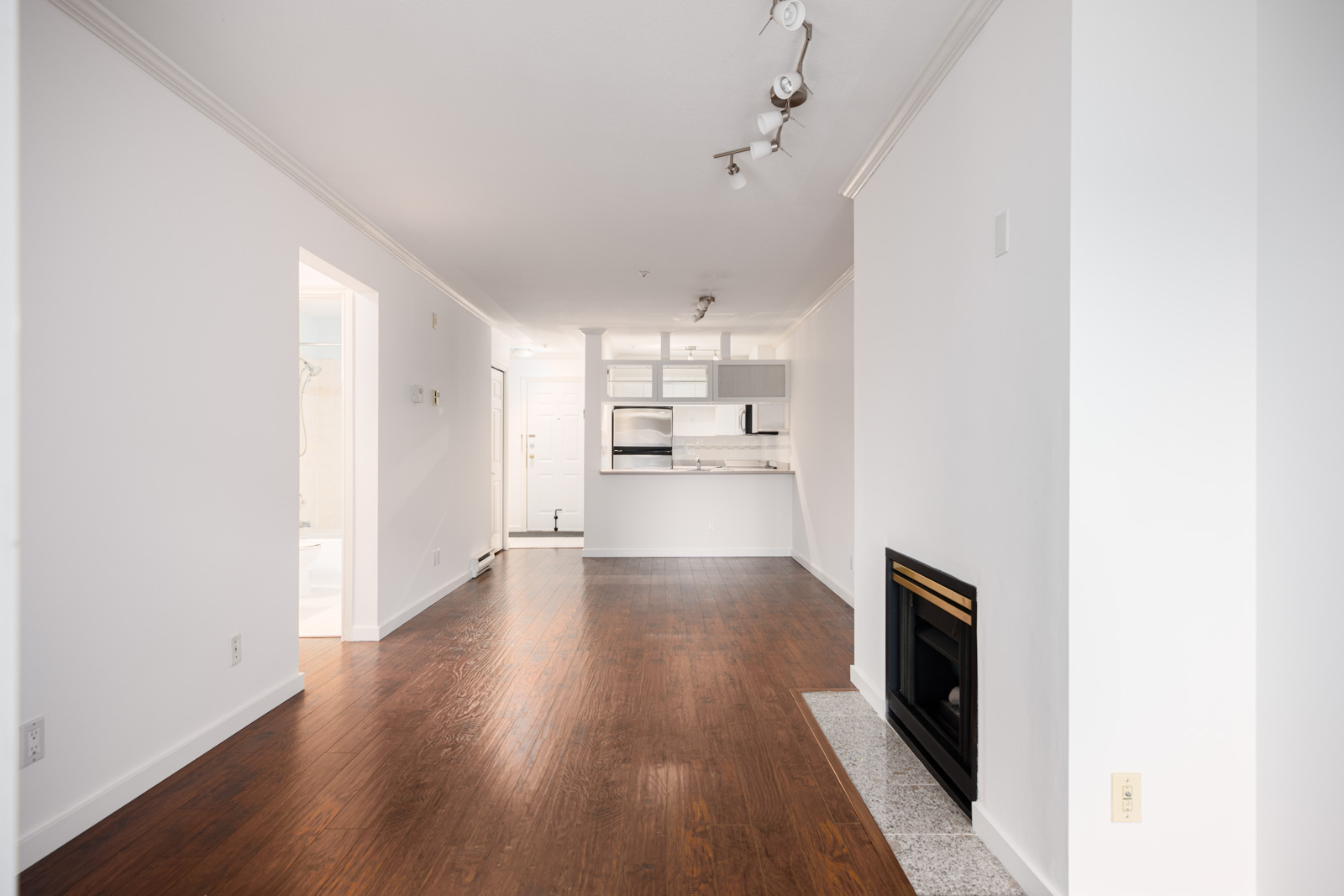 Empty apartment interior with white walls, wooden floors, a fireplace on the right, and a kitchen area with white cabinetry and stainless steel appliances in the background.
