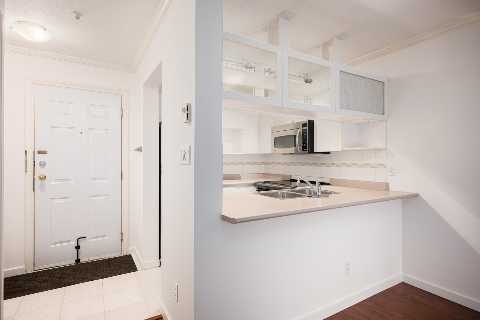 Small kitchen with white cabinets, built-in microwave, and a light-colored countertop next to an entrance door with a tiled entryway and coat hook.