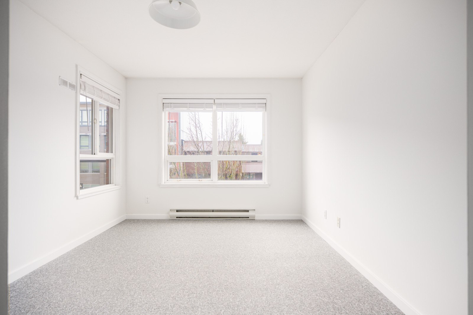 Empty, unfurnished white room with gray carpet, large window, and a ceiling light fixture, featuring outdoor view of a building and trees.