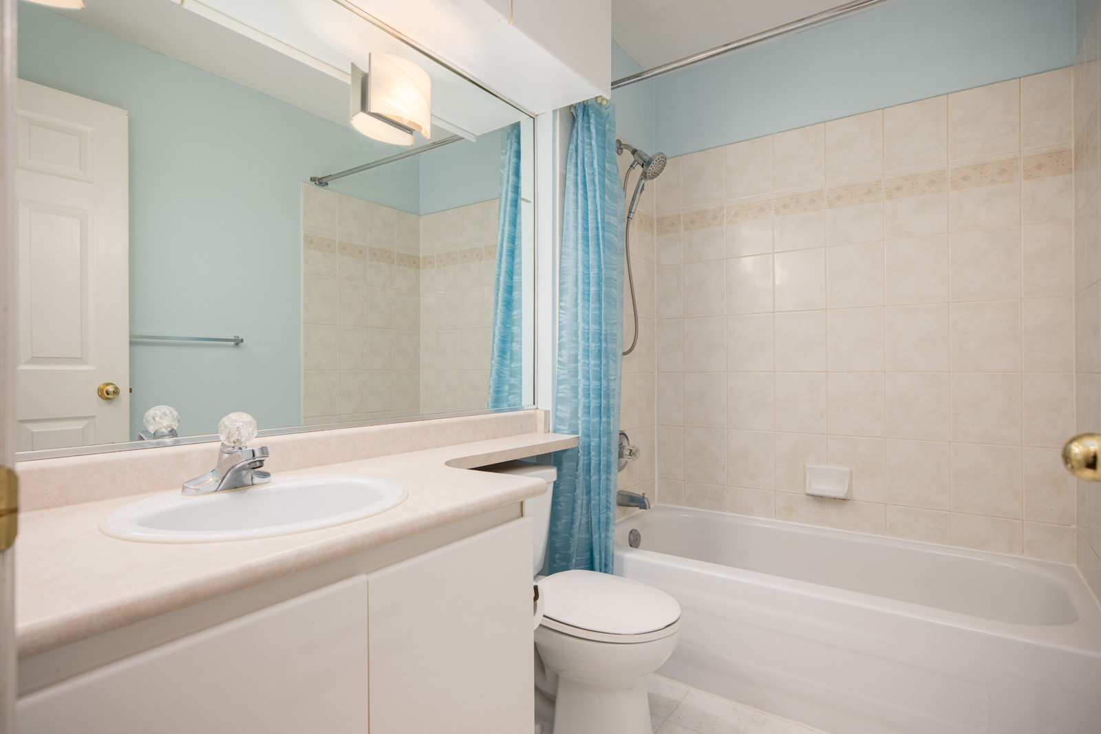A bathroom with a sink, countertop, toilet, bathtub, showerhead, blue shower curtain, and tiled walls. A mirror and towel rack are also visible.