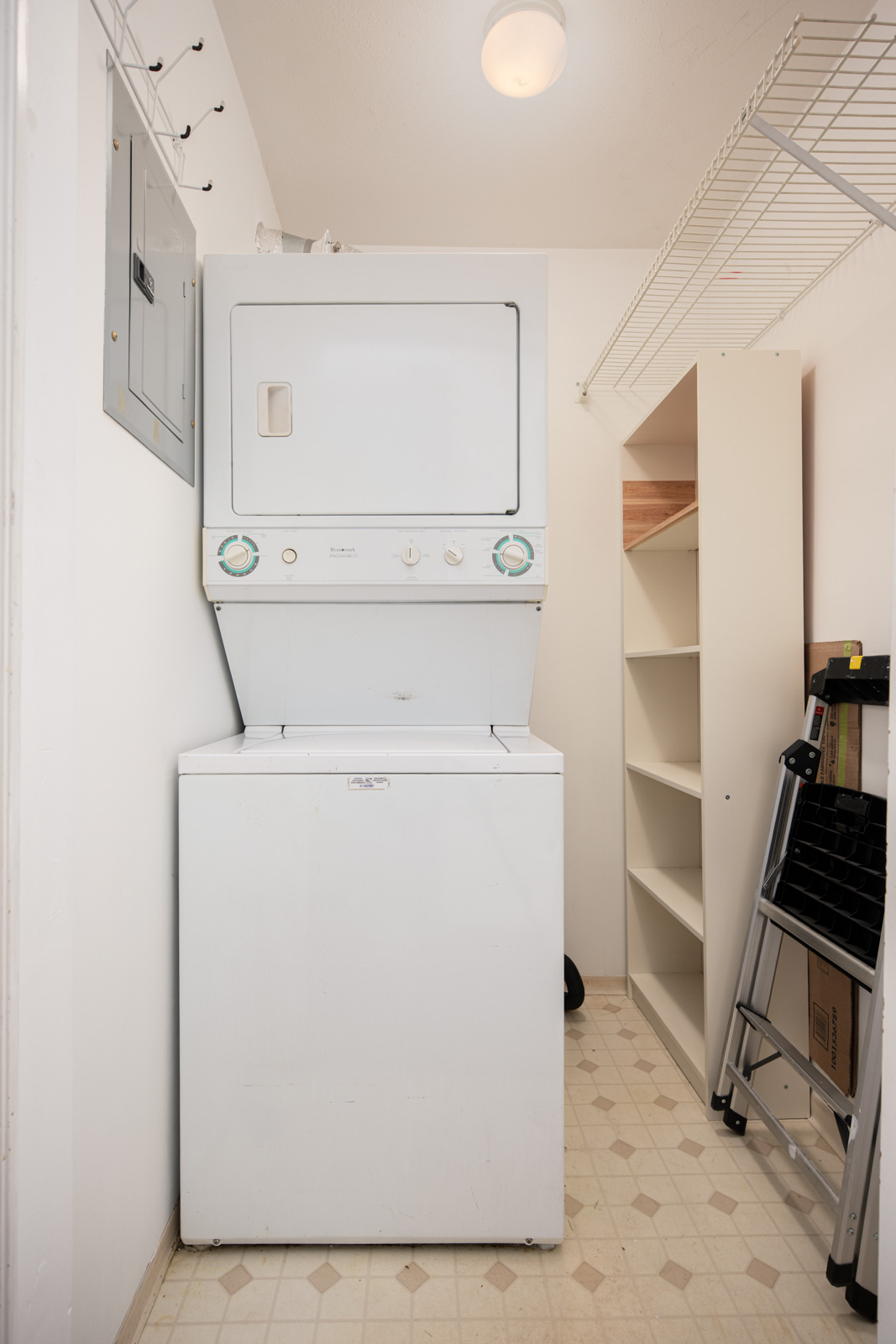 A stacked washer and dryer unit in a small laundry room with white walls, storage shelves, a folding step ladder, and overhead wire rack.