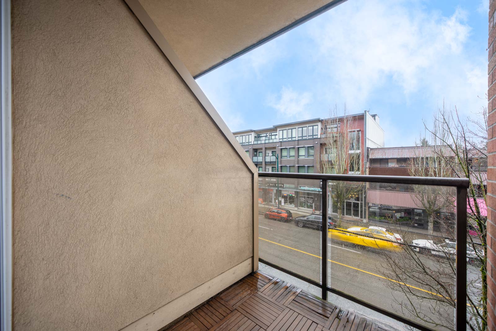 Small balcony with wood flooring, glass railing, and beige wall, overlooking a street with parked cars and commercial buildings on the opposite side.