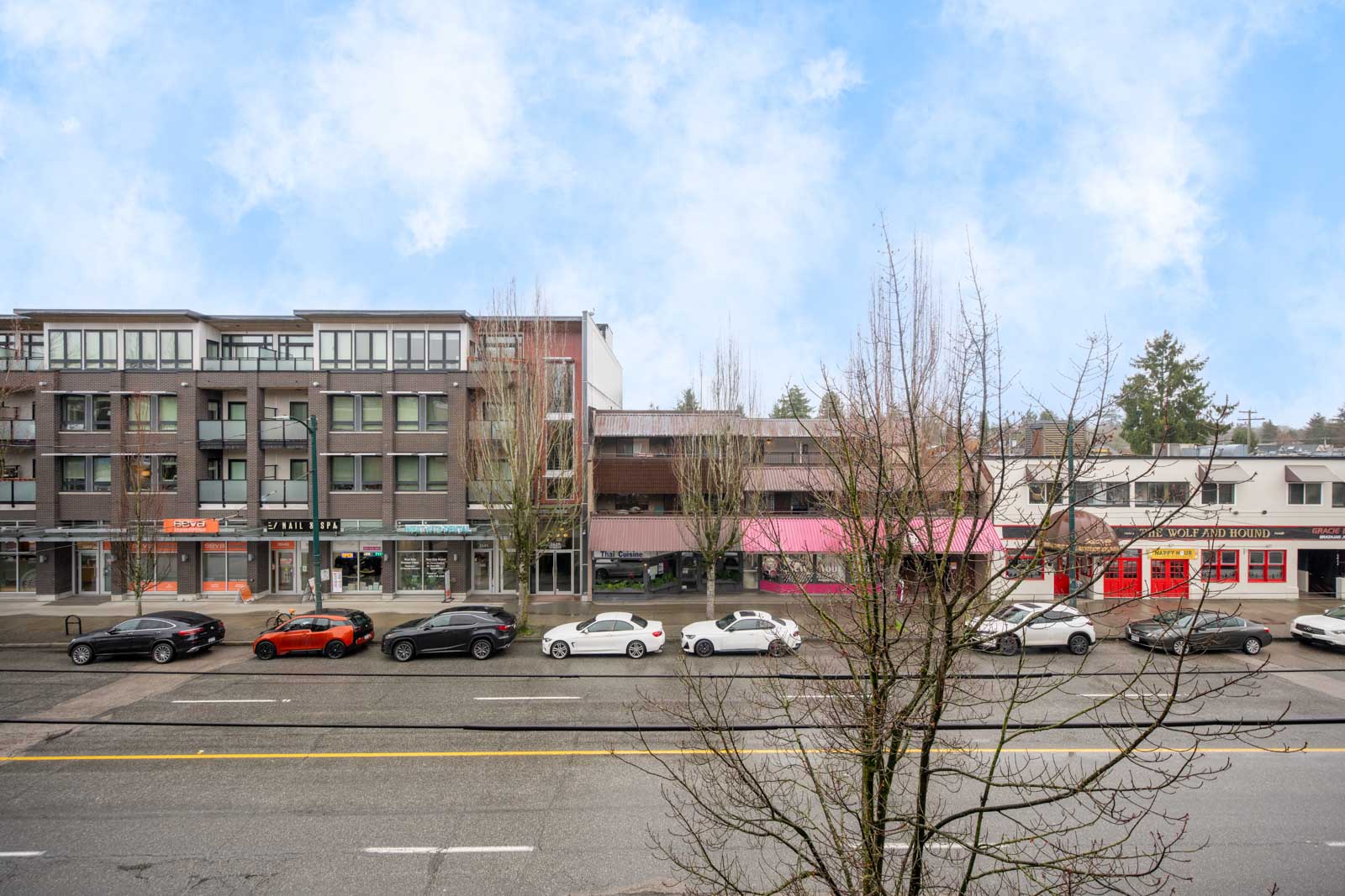 A city street with parked cars, leafless trees, and low-rise buildings containing shops and businesses under a partly cloudy sky.