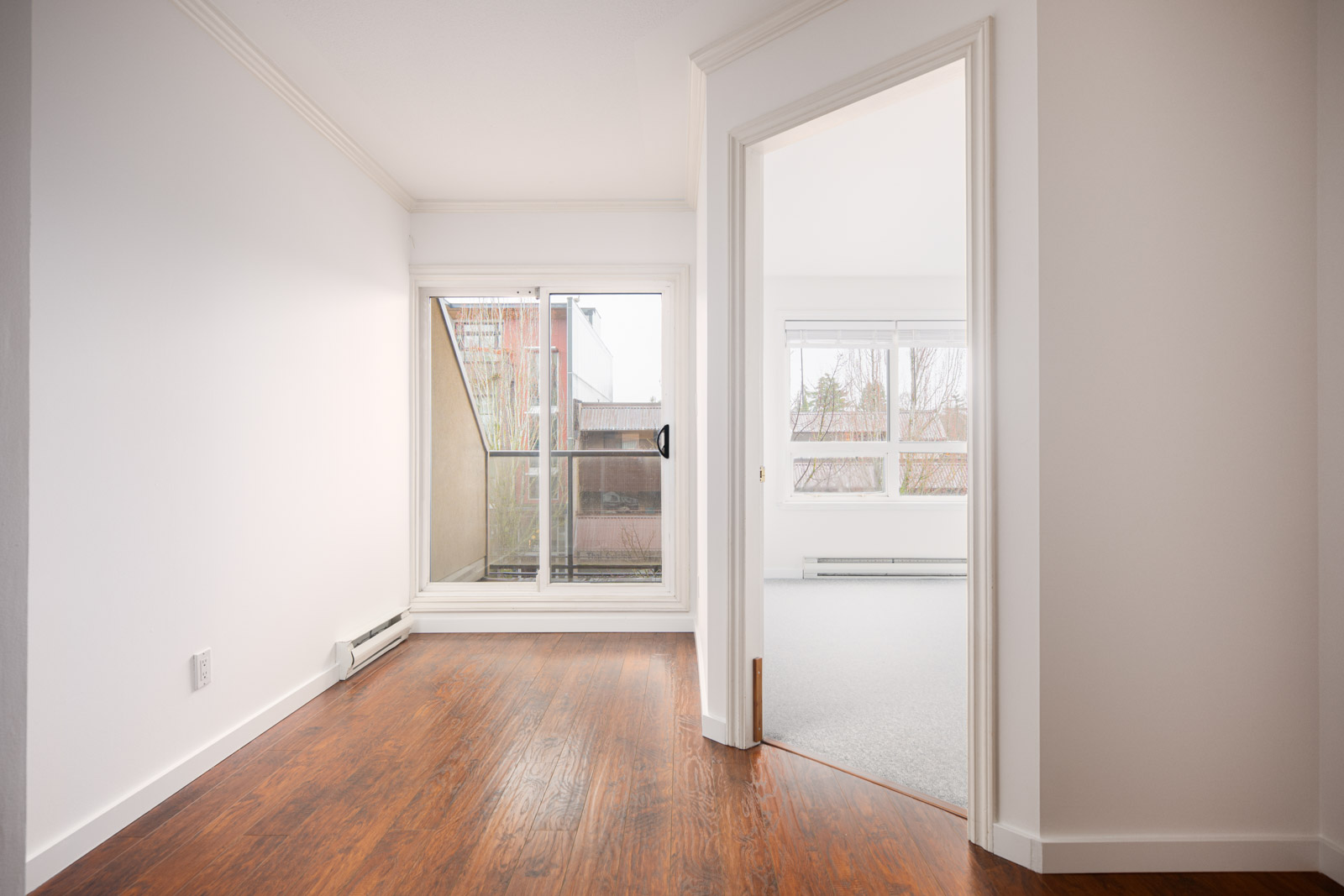 A bright apartment interior with hardwood flooring, a sliding glass door leading to a small balcony, and an adjacent carpeted room with large windows.