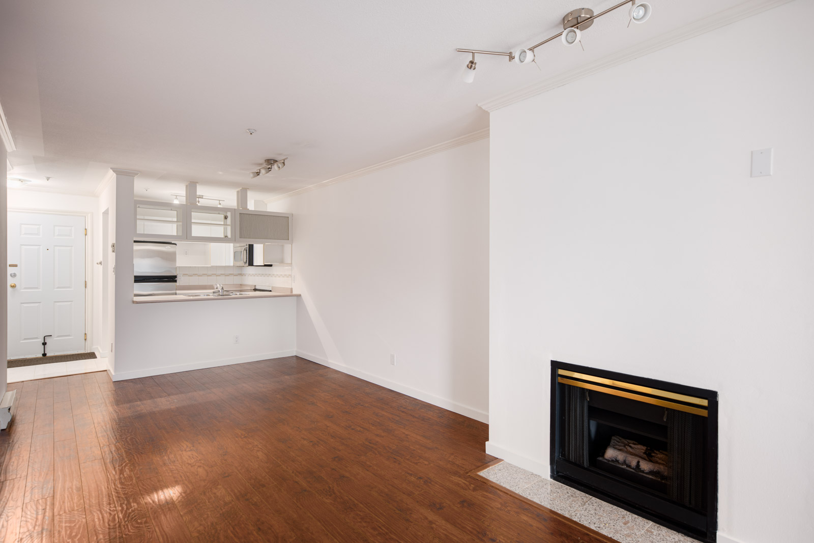 A modern, unfurnished living area with wood flooring, a fireplace, white walls, and an open kitchen with white cabinets and countertops in the background.