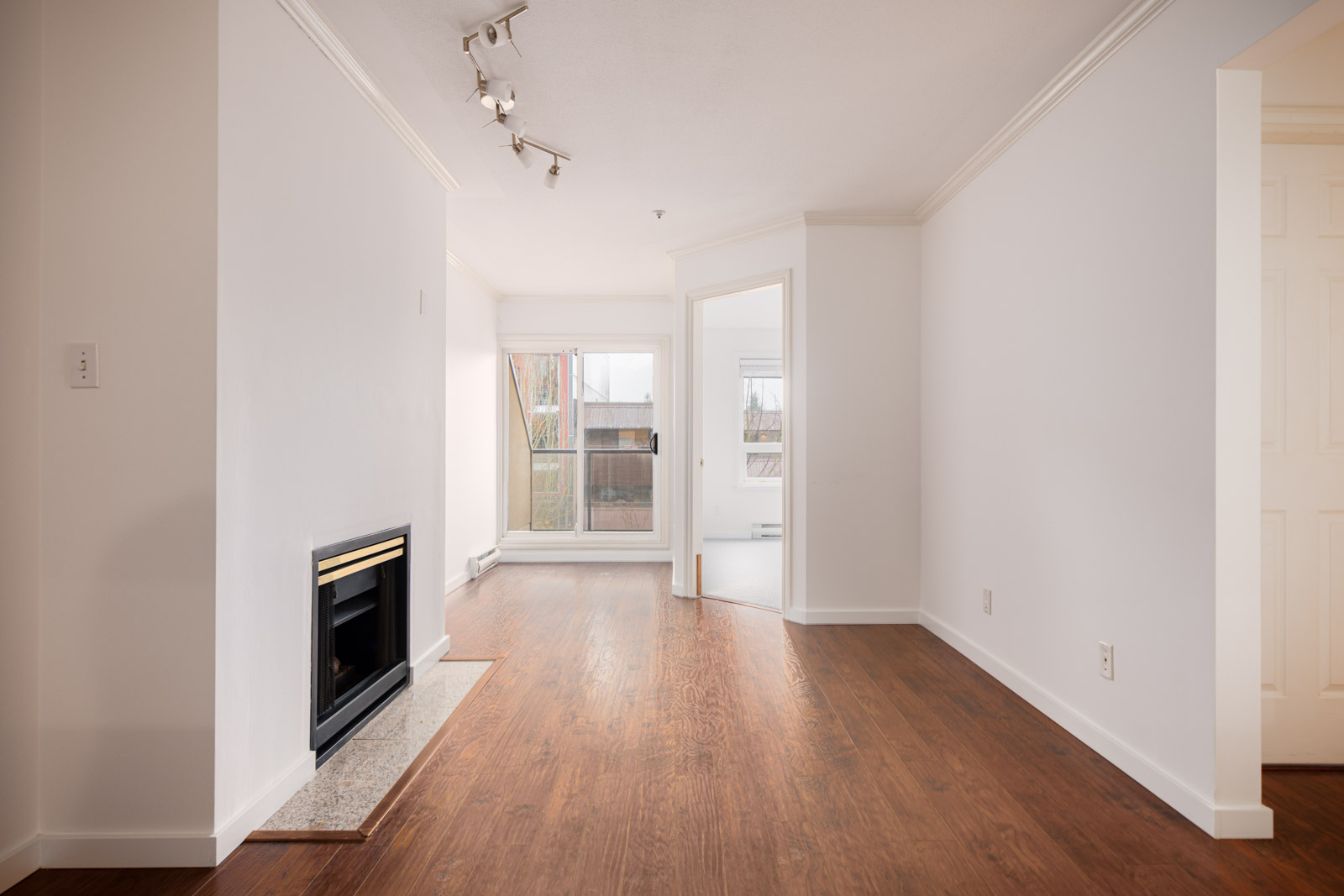 Unfurnished living room with wood flooring, a fireplace on the left, white walls, ceiling lights, and large windows letting in natural light.
