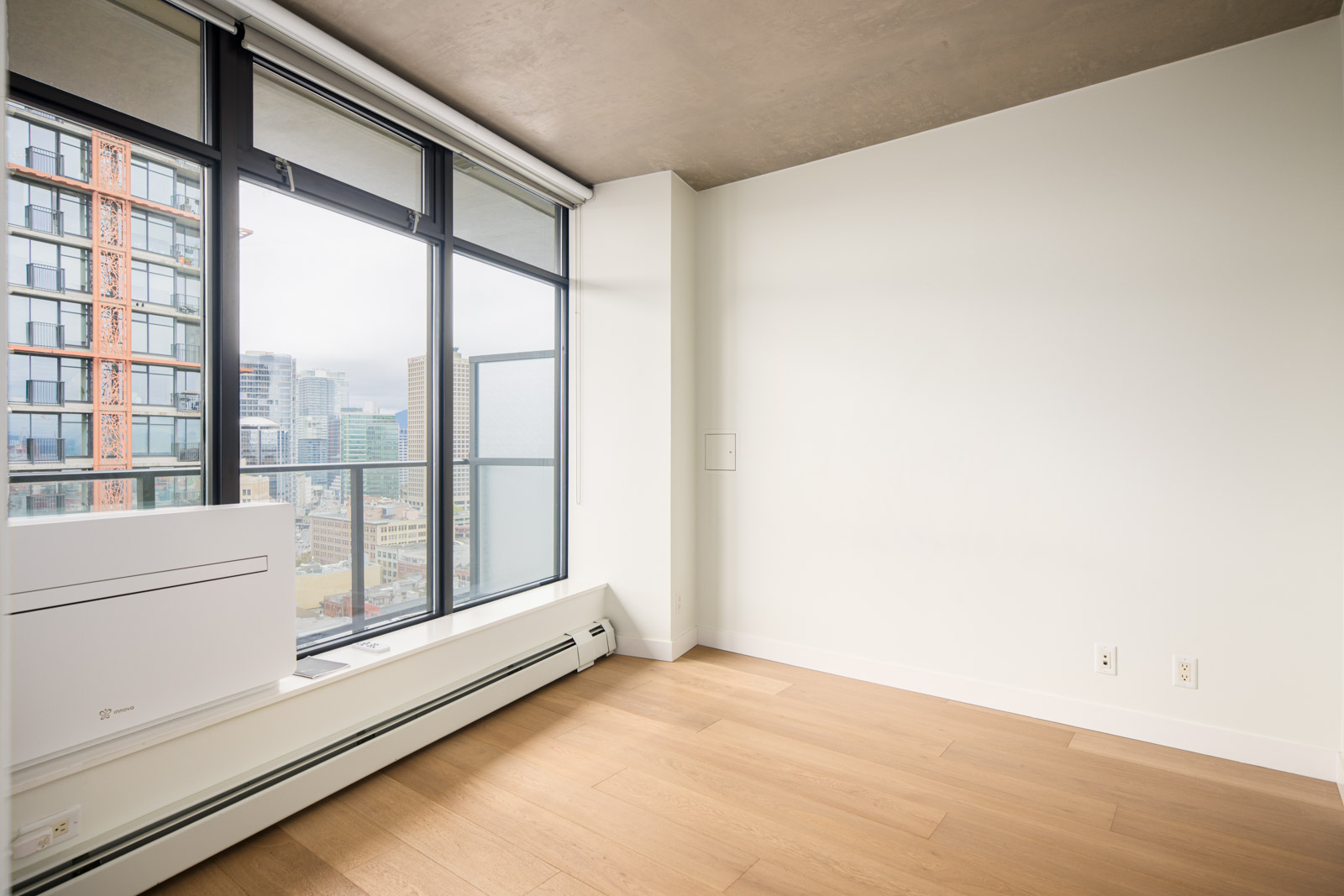 Unfurnished modern apartment room with light wood flooring, large floor-to-ceiling windows, and a view of city buildings.