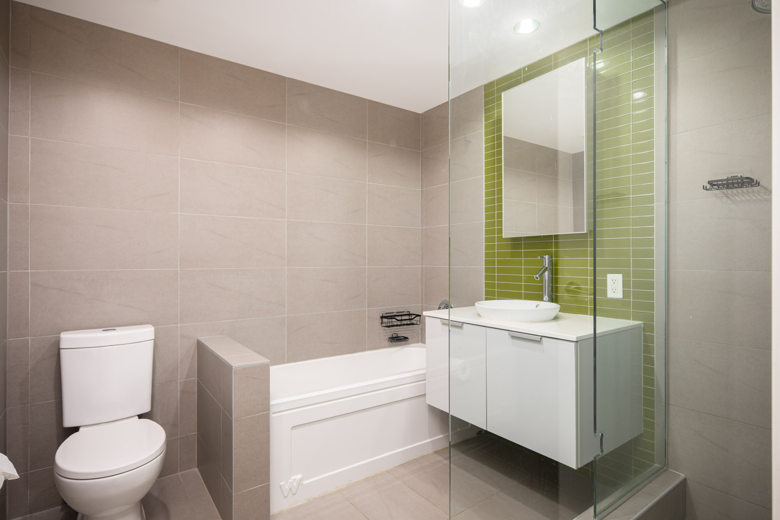 Modern bathroom with a toilet, bathtub, glass-enclosed shower, wall-mounted sink cabinet, and green tile accent wall behind a mirror.
