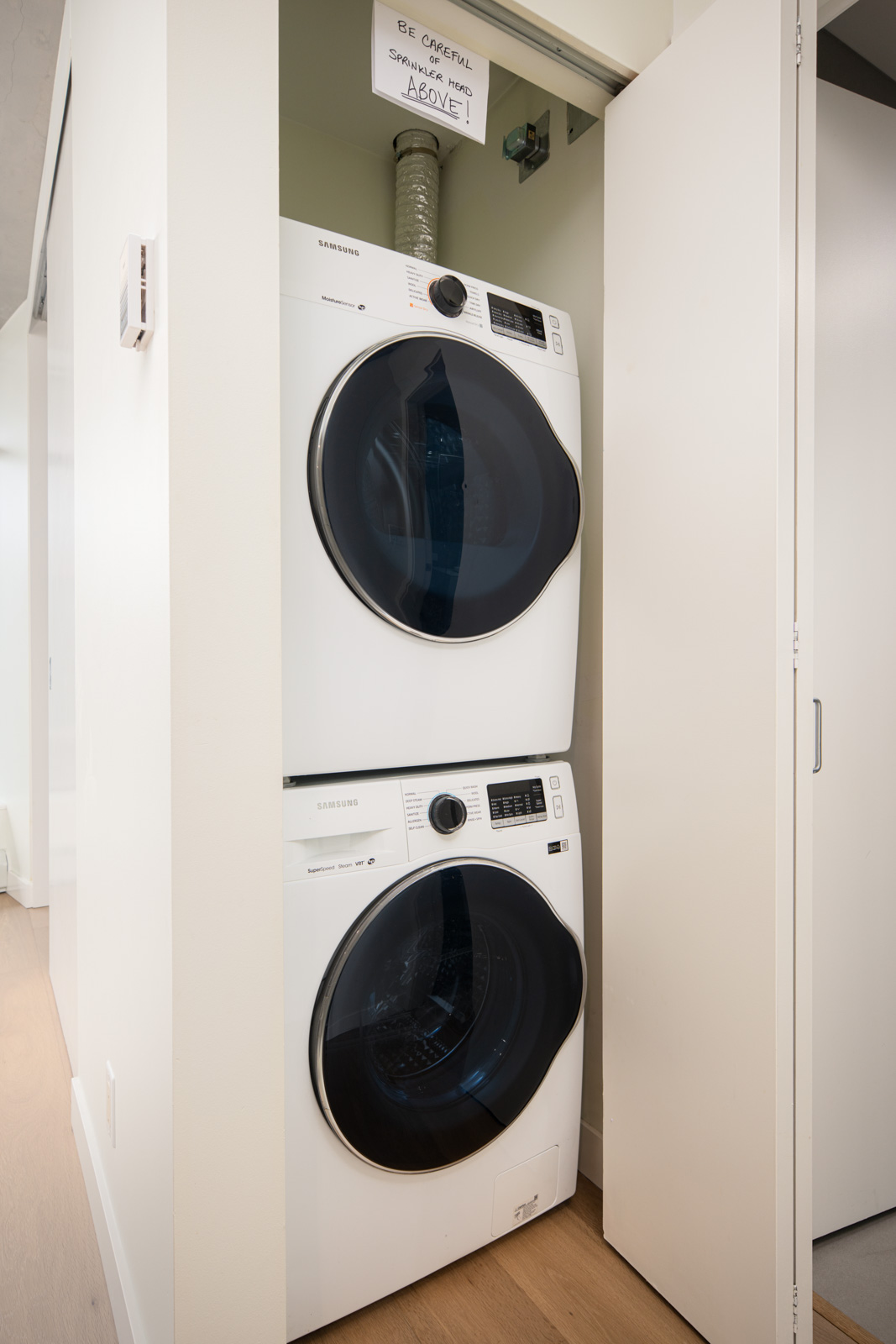A stacked washer and dryer unit is installed in a small closet. A handwritten sign above warns to be careful of the exposed vent pipe.