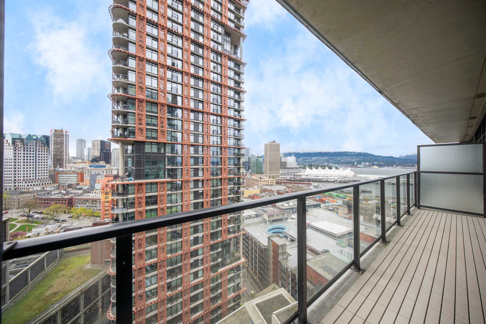 View from a balcony with glass railing overlooking a cityscape, featuring a tall red building and distant waterfront structures under a partly cloudy sky.