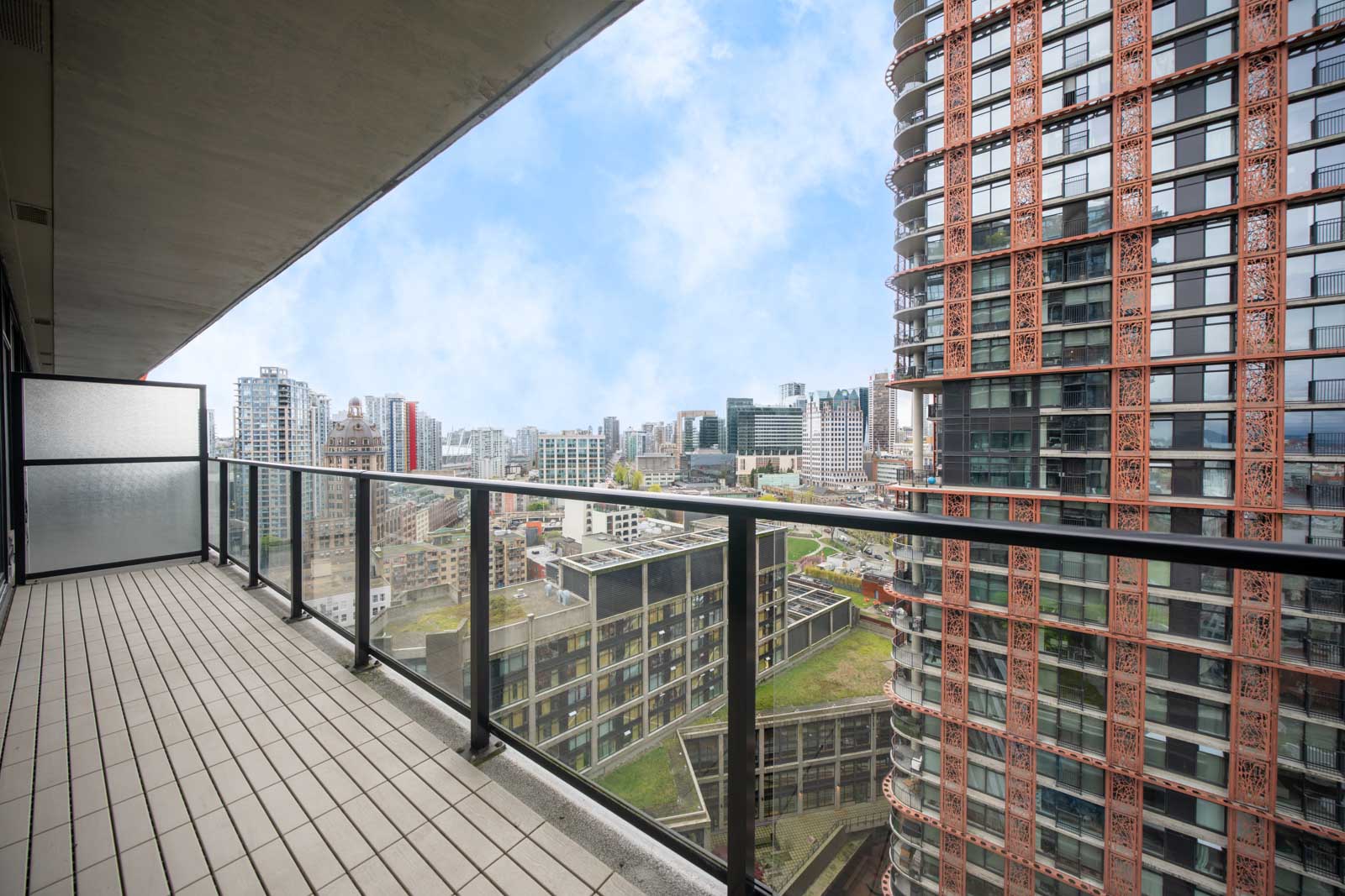 High-rise apartment balcony with glass railing, overlooking modern city buildings under a partly cloudy sky.