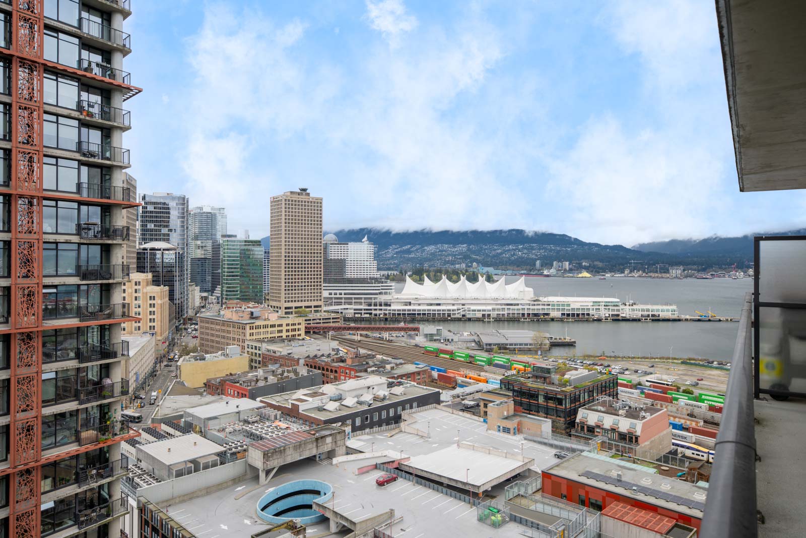A high-angle view of a cityscape with tall buildings, a waterfront convention center, a rail yard with colorful containers, and mountains in the background under a partly cloudy sky.