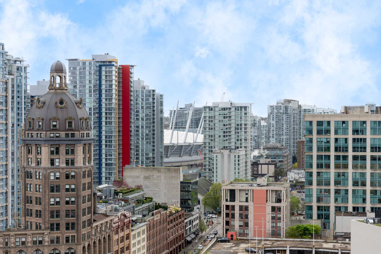 A cityscape view featuring modern high-rise buildings, an older domed building in the foreground, and a stadium with white roof supports in the background under a partly cloudy sky.