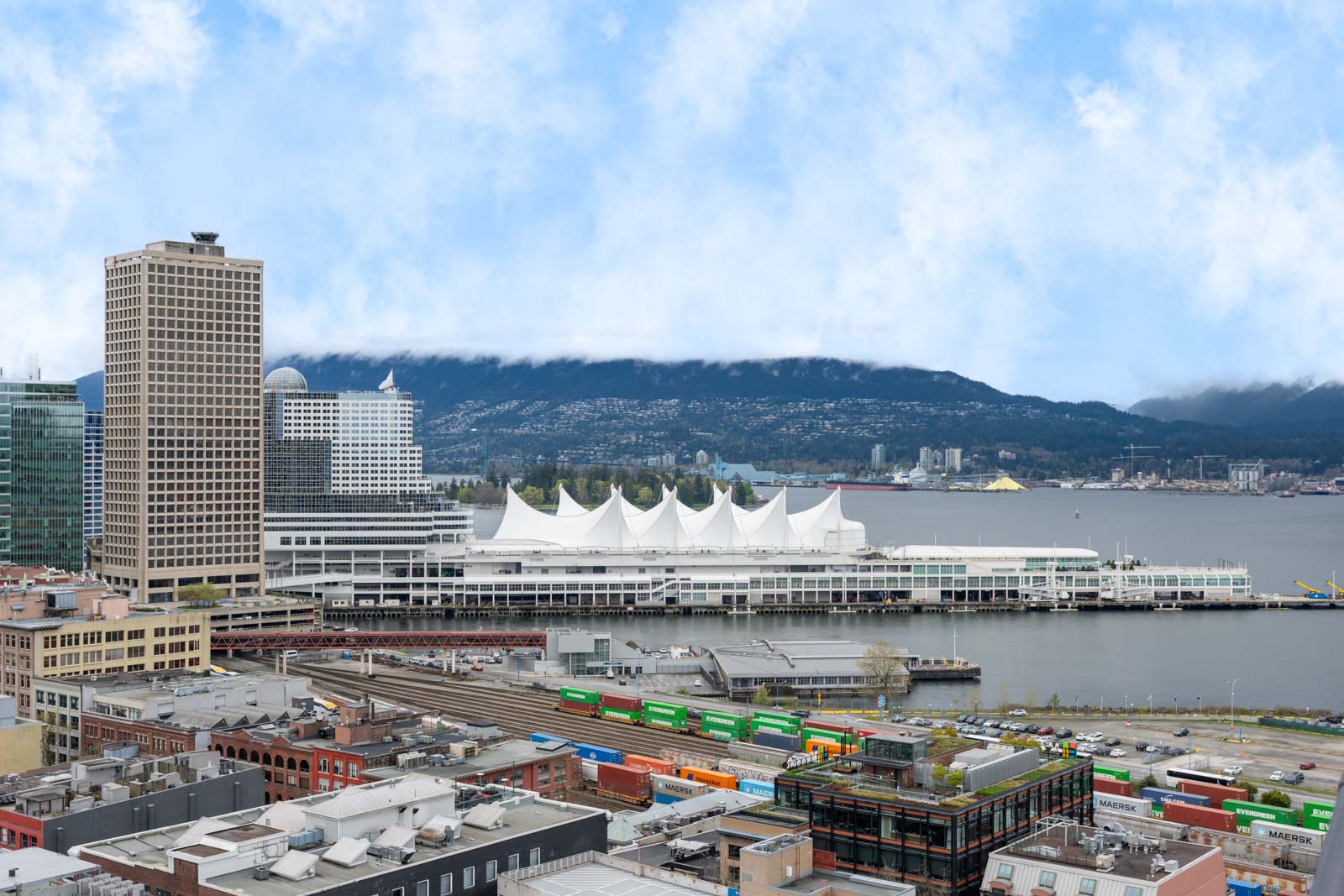 A waterfront cityscape with a white tent-roofed building, high-rise towers, railway tracks, and colorful shipping containers, set against mountains and a cloudy sky.