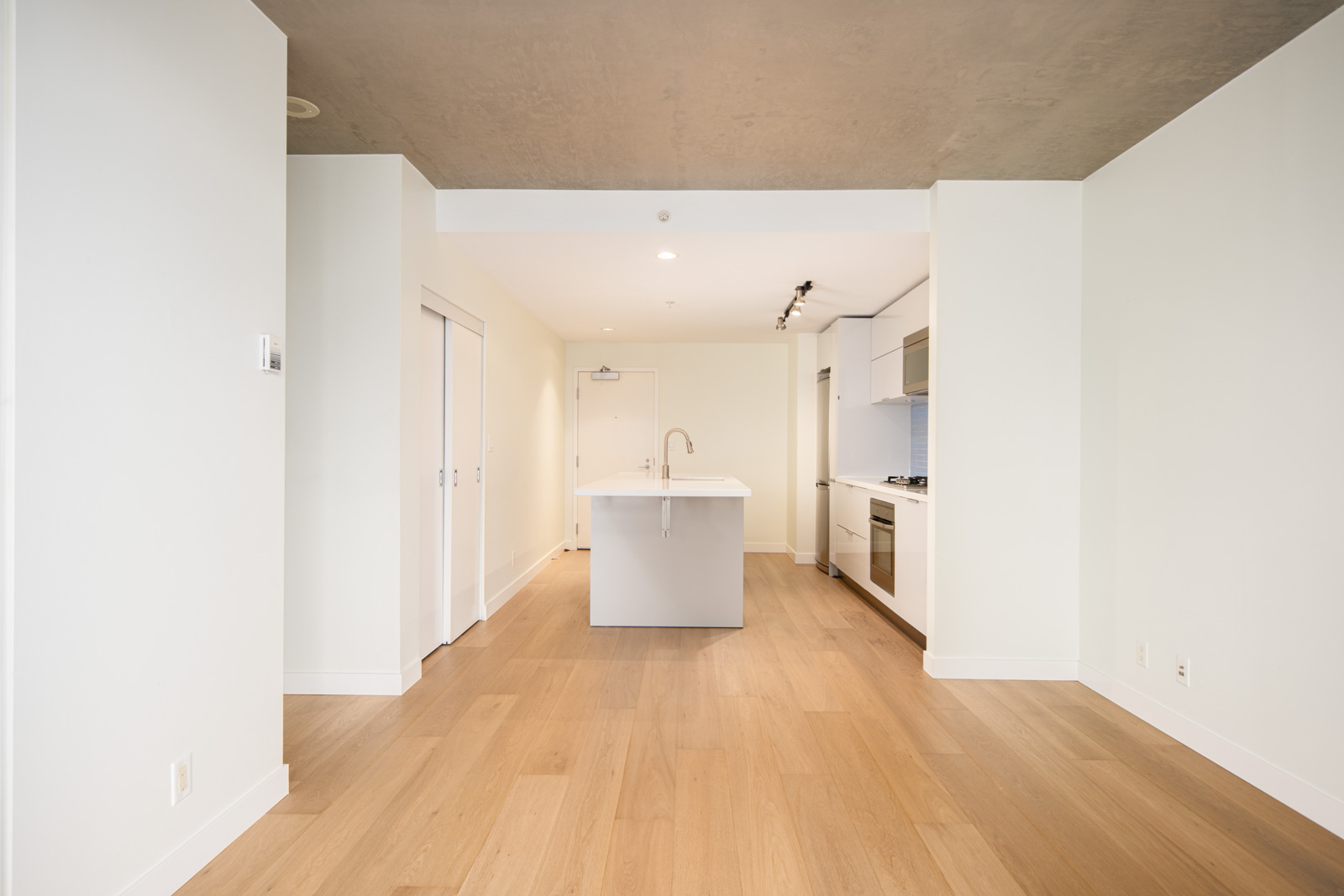 Modern, unfurnished kitchen and living area with light wood flooring, white walls, a central island, and built-in appliances under a gray ceiling.