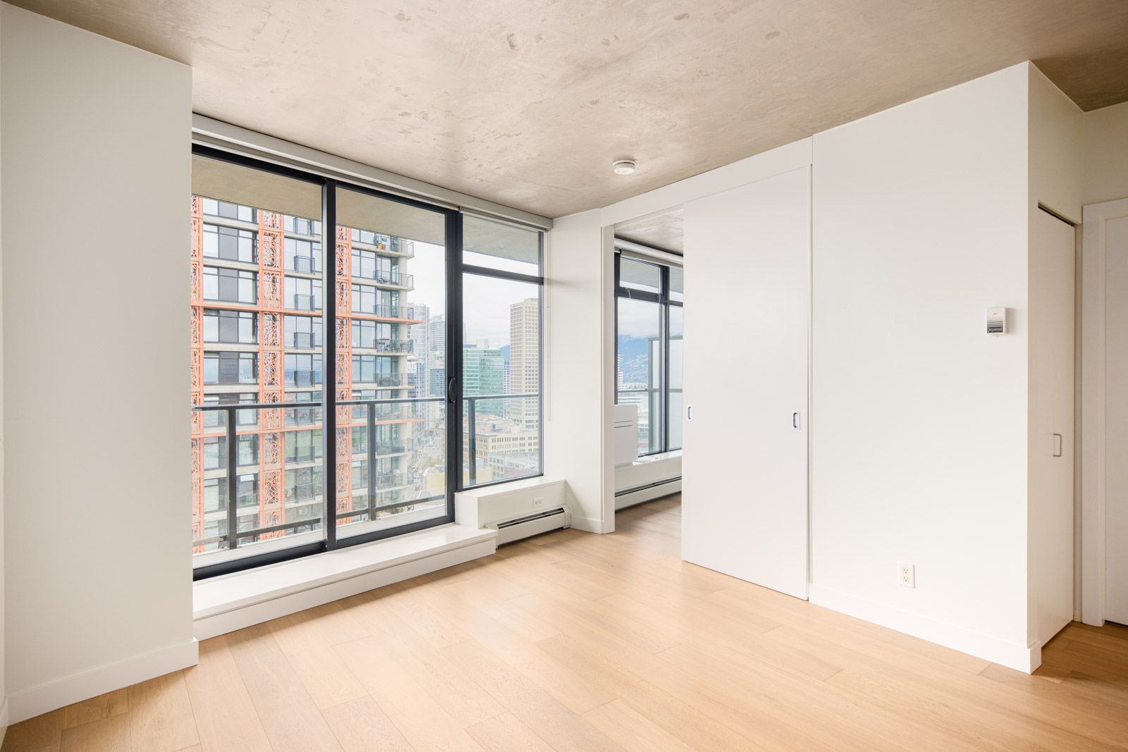 Unfurnished modern apartment room with large floor-to-ceiling windows, light wood flooring, white walls, and a view of city buildings outside.