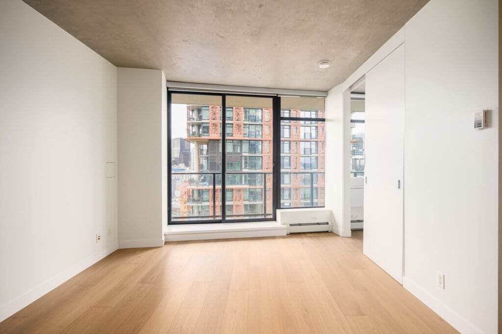 Unfurnished modern apartment room with light wood flooring, large floor-to-ceiling window, and a view of nearby city buildings.