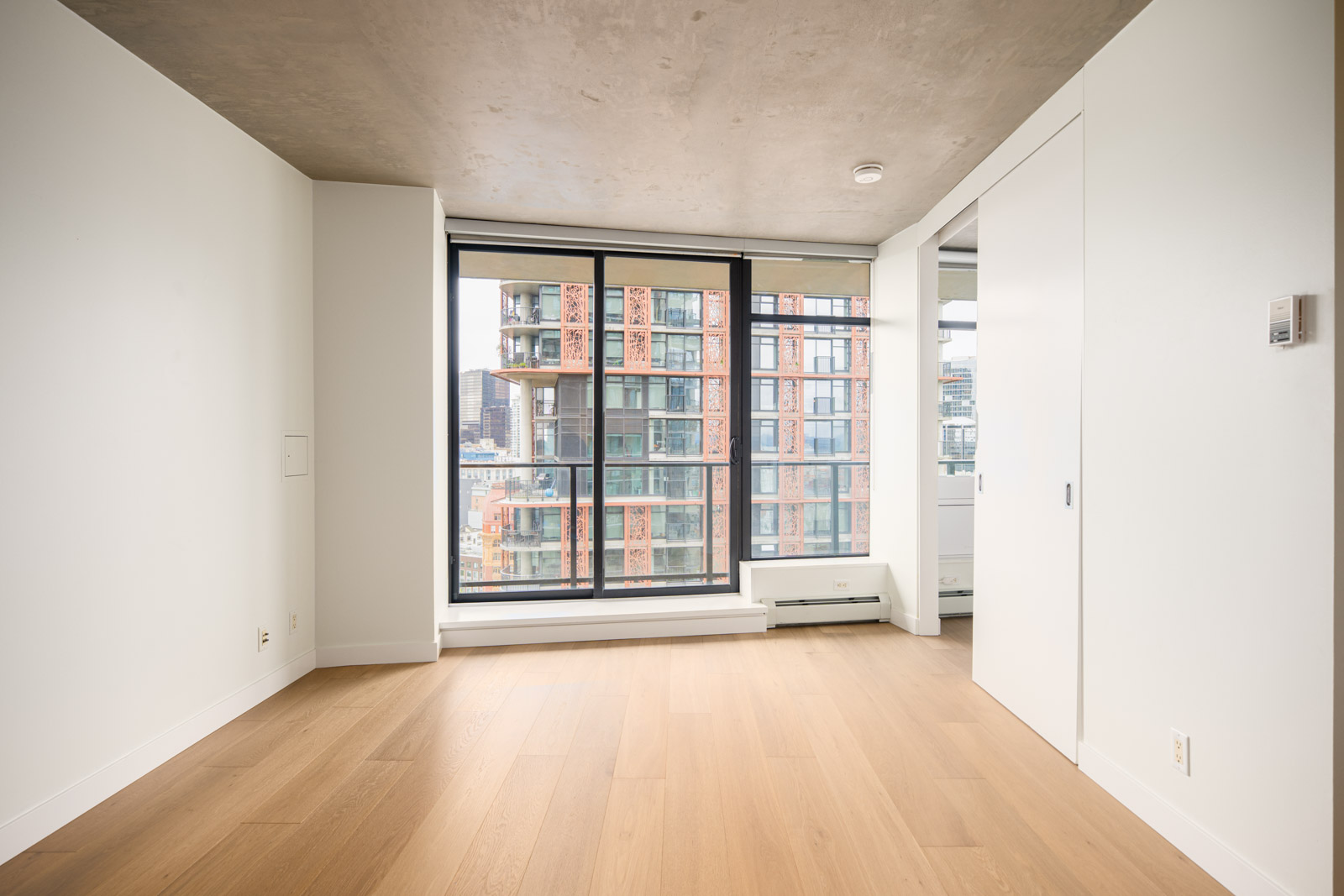Unfurnished modern apartment room with light wood flooring, large floor-to-ceiling window, and a view of nearby city buildings.