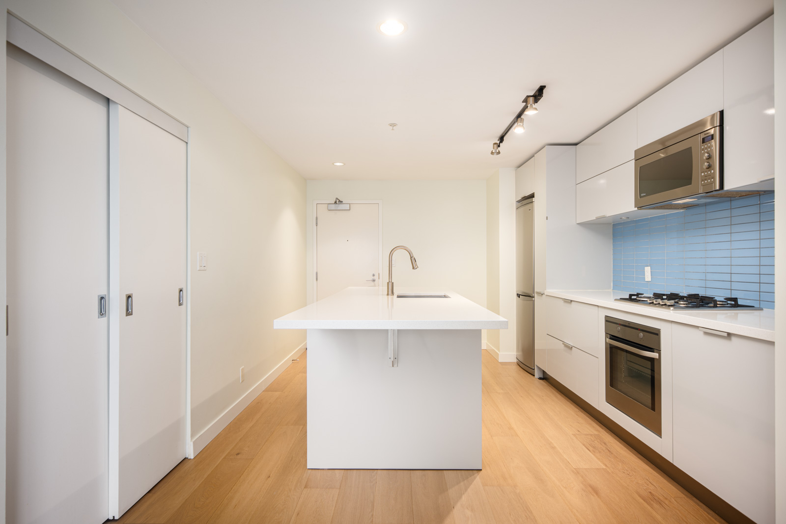 Modern white kitchen with wood flooring, an island with sink, built-in appliances, and blue tile backsplash. Sliding doors are on the left side of the room.