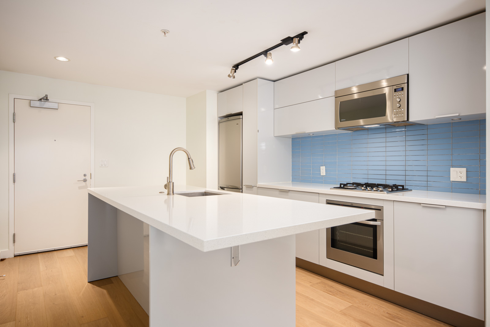 Modern kitchen with white cabinets, island with sink, stainless steel appliances, and blue subway tile backsplash on wood flooring.
