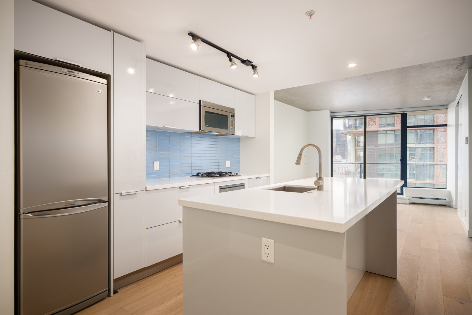 Modern kitchen with white cabinetry, stainless steel appliances, island with sink, and blue tile backsplash; large windows in the background provide natural light.