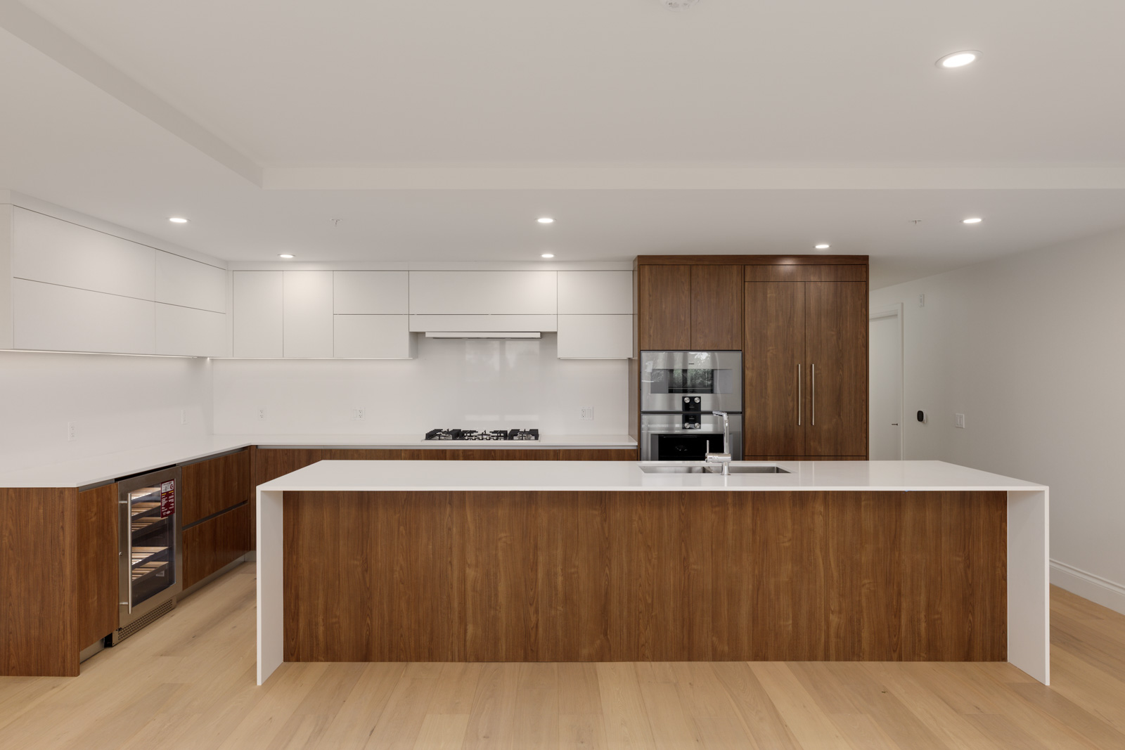 Modern kitchen with white countertops, wood cabinets, built-in oven, stovetop, integrated refrigerator, and a large island with a sink. The floor is light wood, and lighting is recessed.