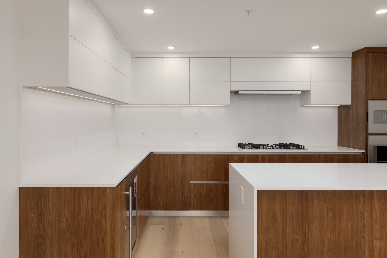 Modern kitchen with white upper cabinets, wood lower cabinets, white countertops, a built-in stove, and integrated oven, all illuminated by recessed ceiling lights.