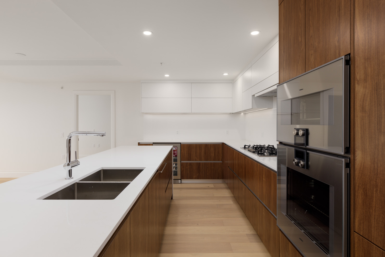 Modern kitchen with wood cabinets, white countertops, built-in oven, gas stove, stainless steel sink, and ample overhead lighting. The space is clean and uncluttered.