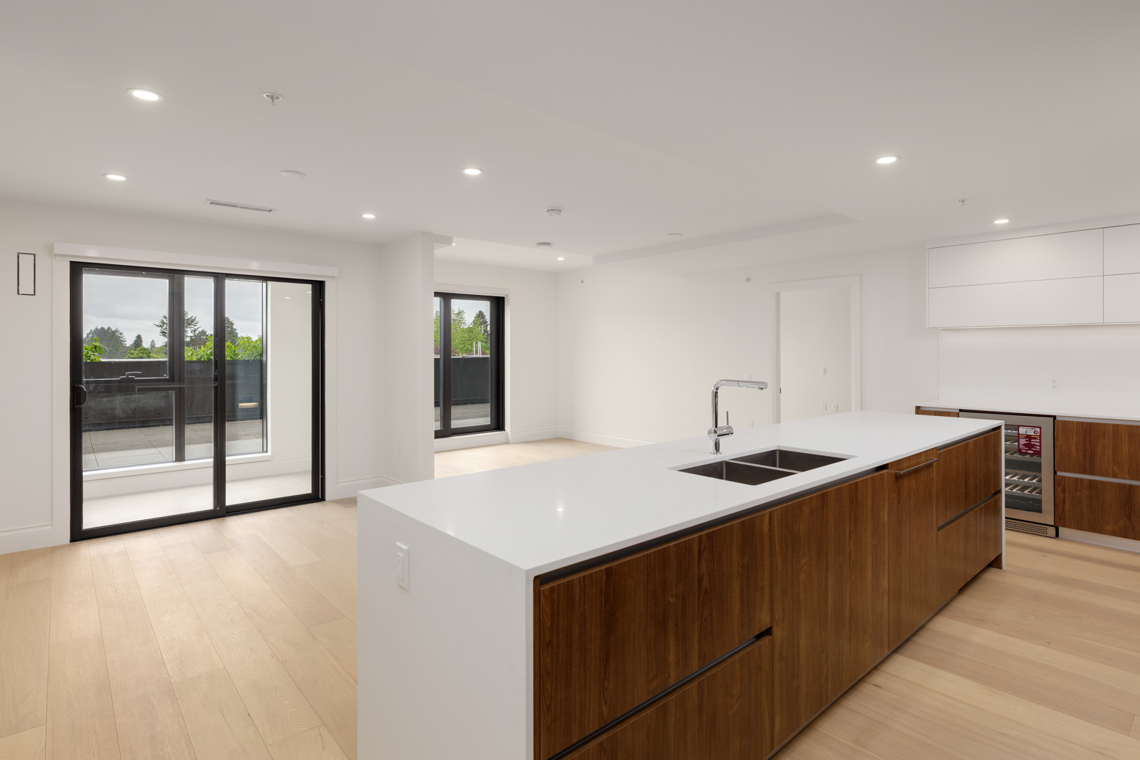 Modern open-plan kitchen and living area with wood flooring, white walls, large island, built-in appliances, and sliding glass doors leading to a balcony.