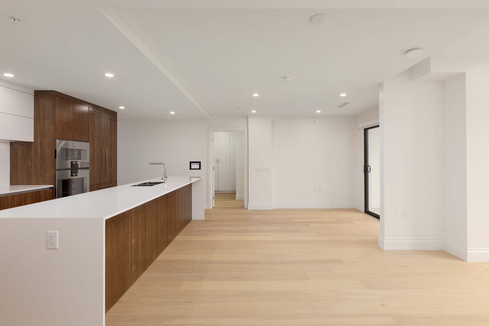 Modern, empty kitchen with wood cabinets, white countertops, built-in appliances, and light wood floors extending into an open living area with sliding glass doors.