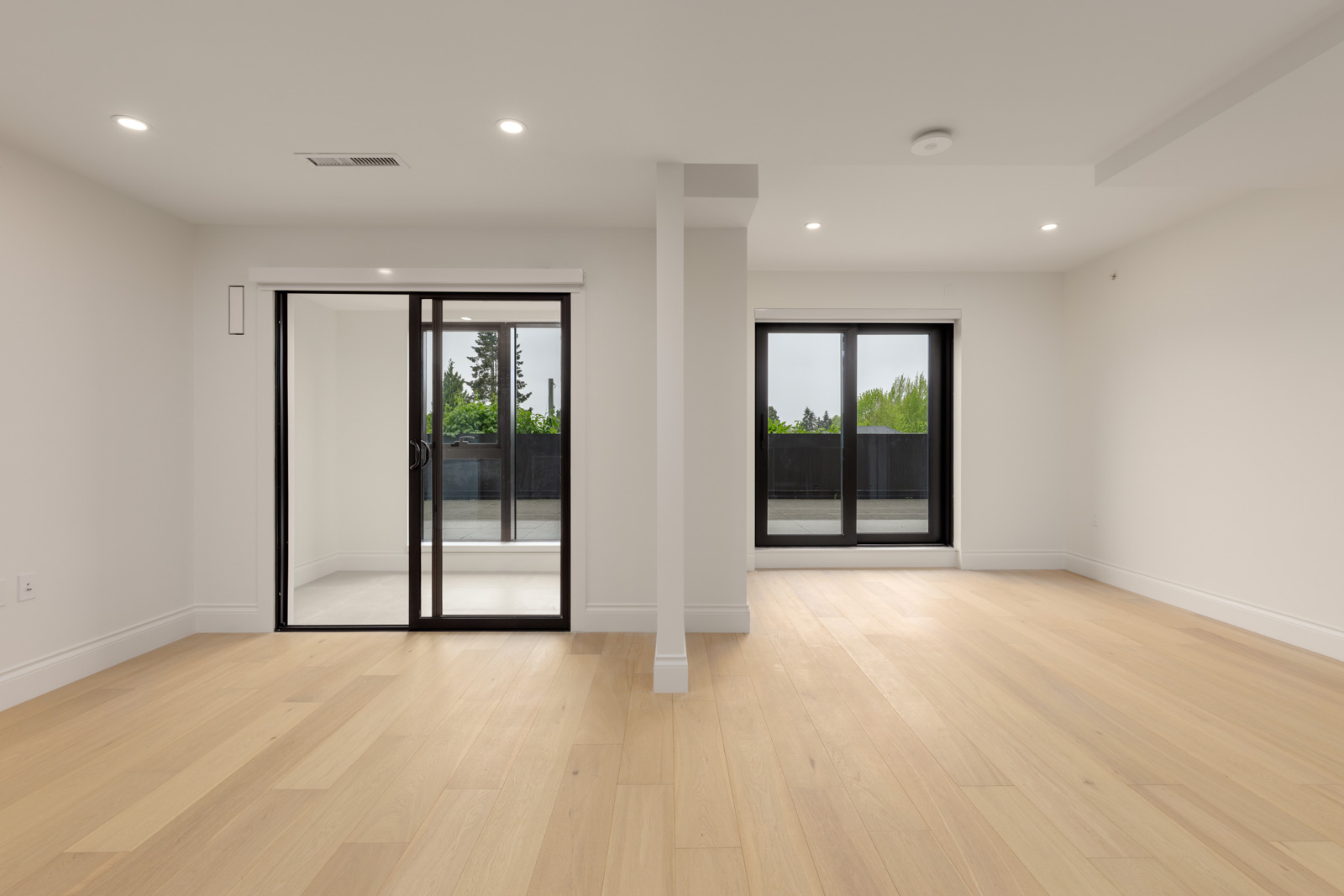 A spacious, empty room with light wood flooring, white walls, recessed lighting, and two large sliding glass doors leading to an outdoor patio or balcony.