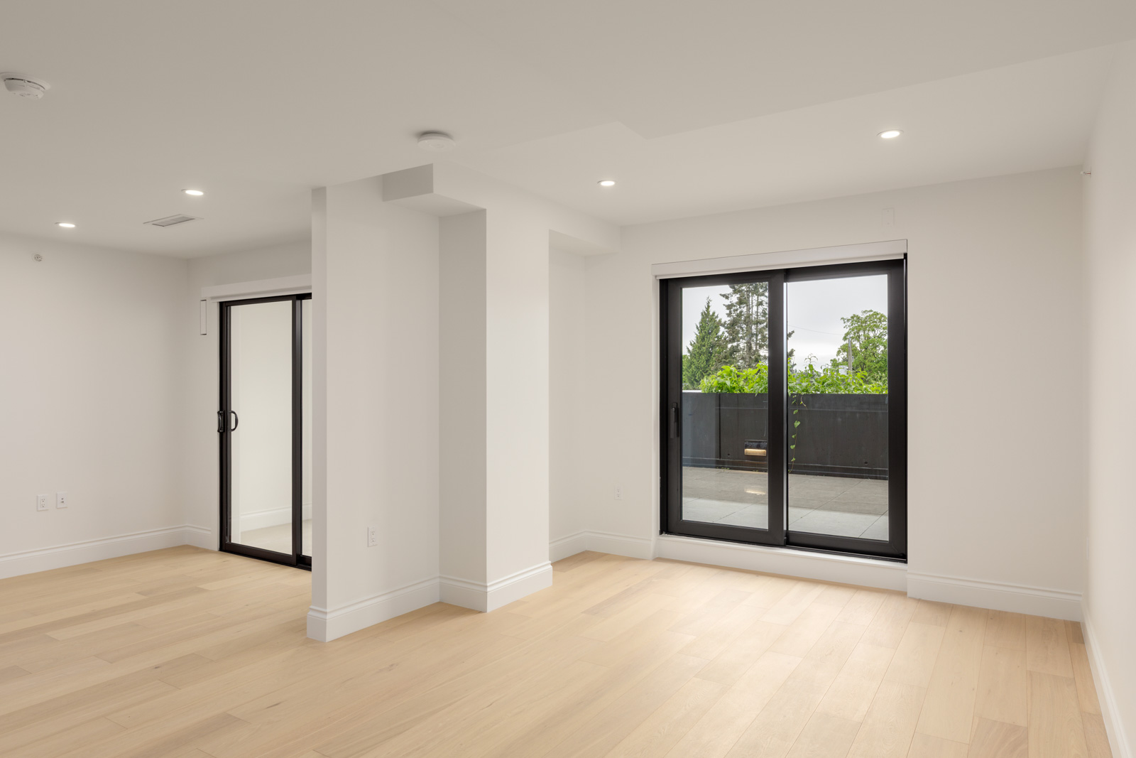 A modern, empty room with light wood floors, white walls, recessed lighting, and large sliding glass doors leading to an outdoor patio with trees visible.