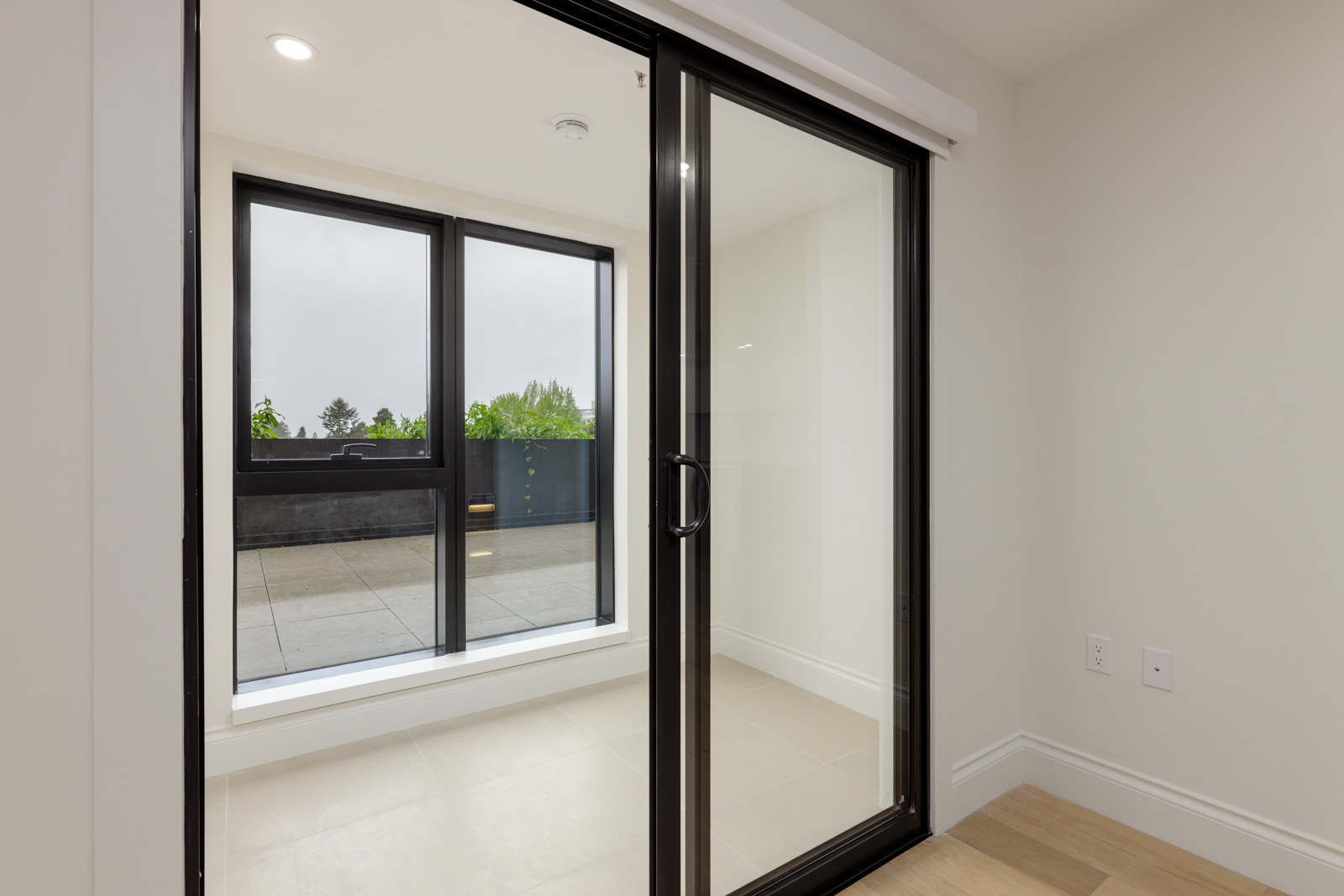 A sliding glass door opens to a small enclosed balcony with tiled flooring and black railing, seen from inside a neutral-colored room.