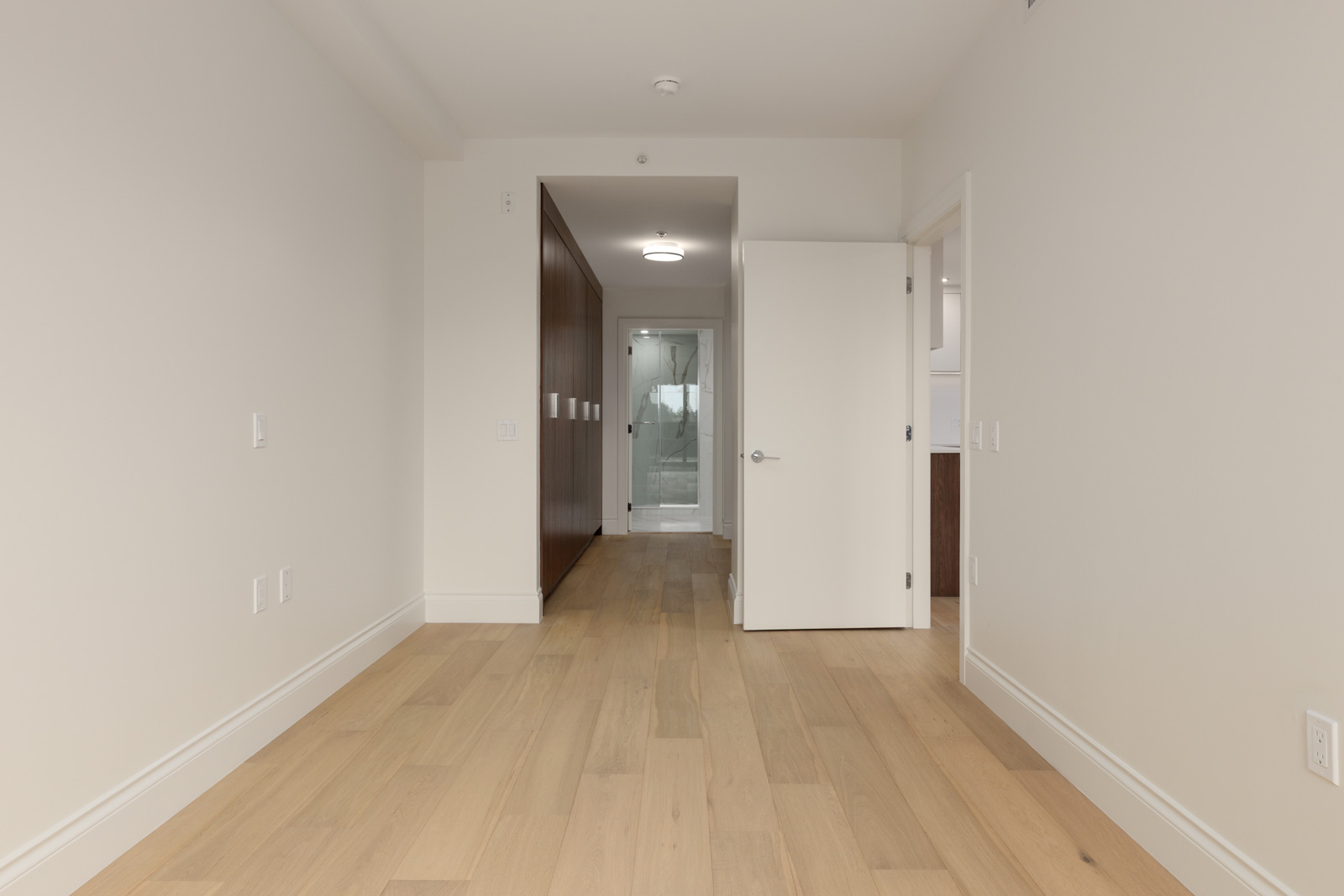 Empty room with light wood flooring, neutral-colored walls, an open white door, and a view into a bathroom with glass shower at the far end.
