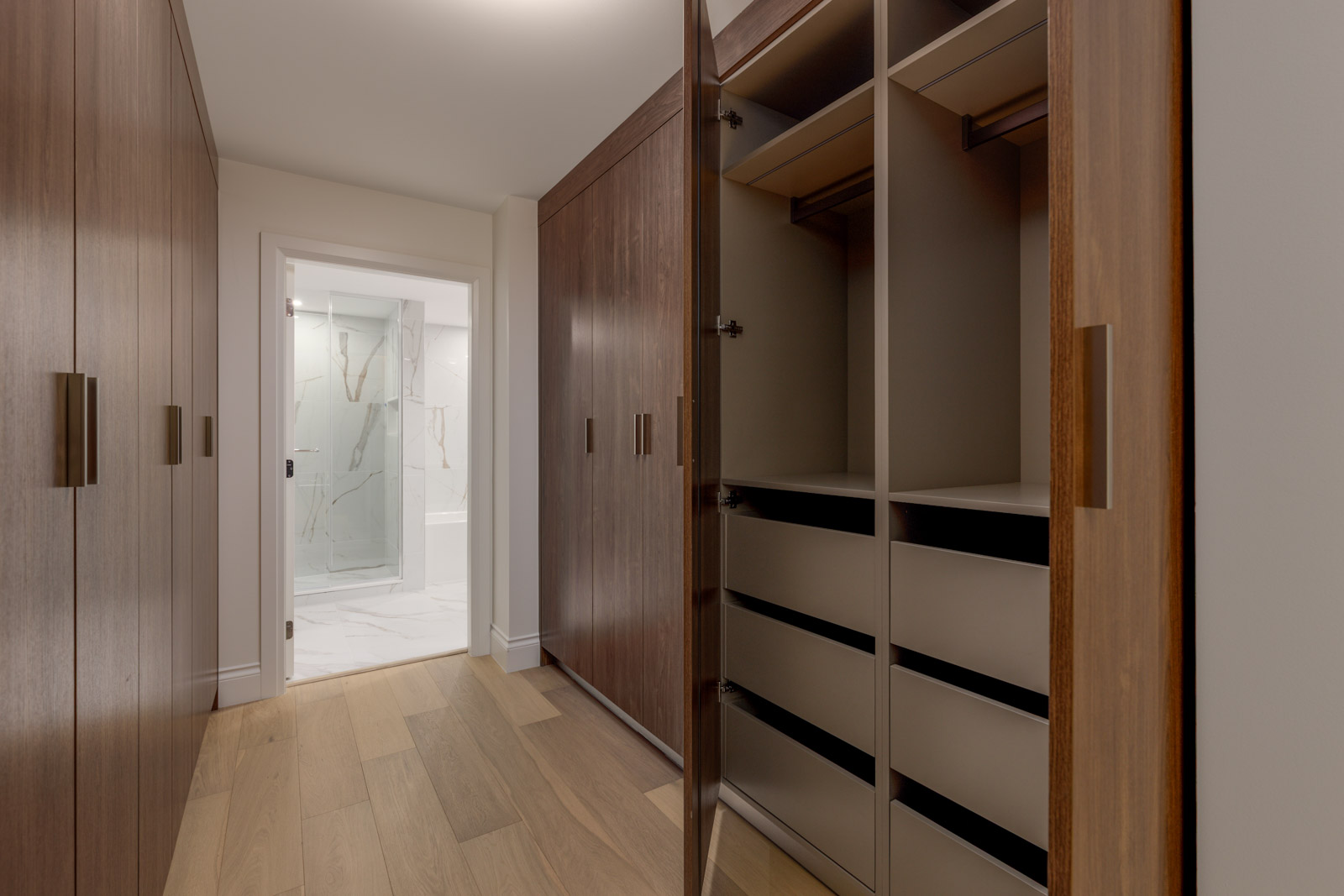 A modern walk-in closet with open wooden cabinets and drawers, light wood flooring, and a view into a bathroom with marble walls.