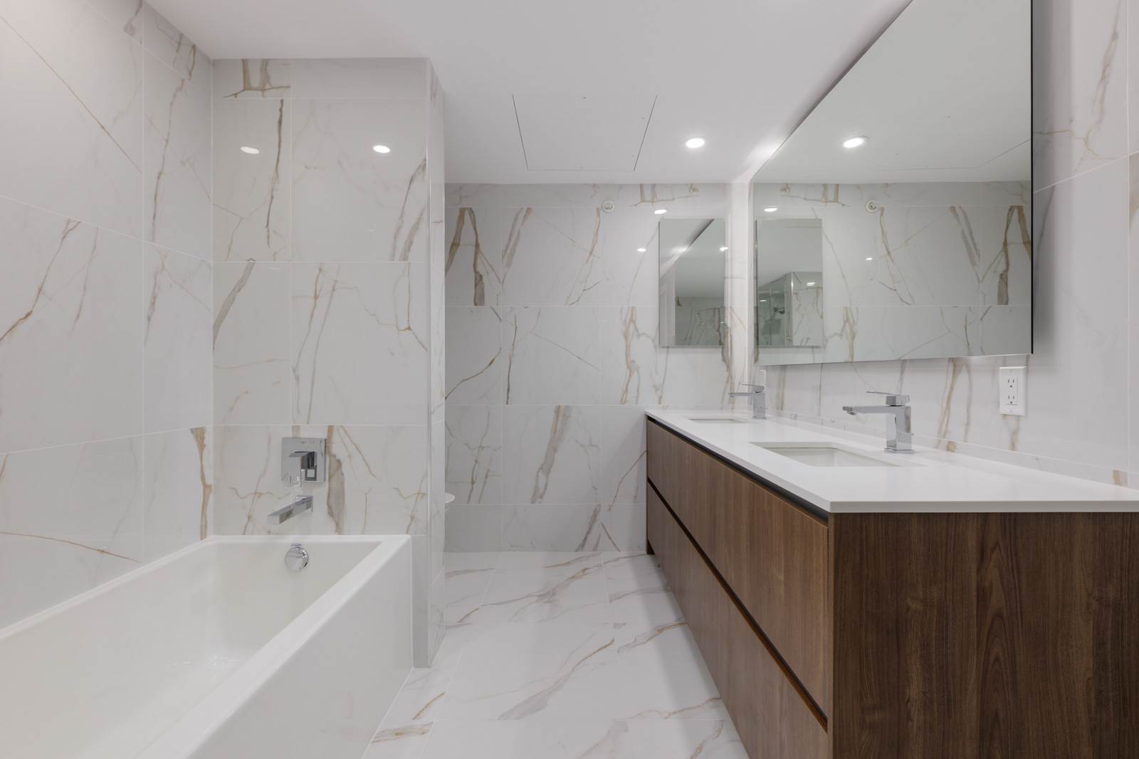 Modern bathroom with white marble walls and floor, a built-in bathtub, a wooden vanity with dual sinks, and a large mirror above the counter.