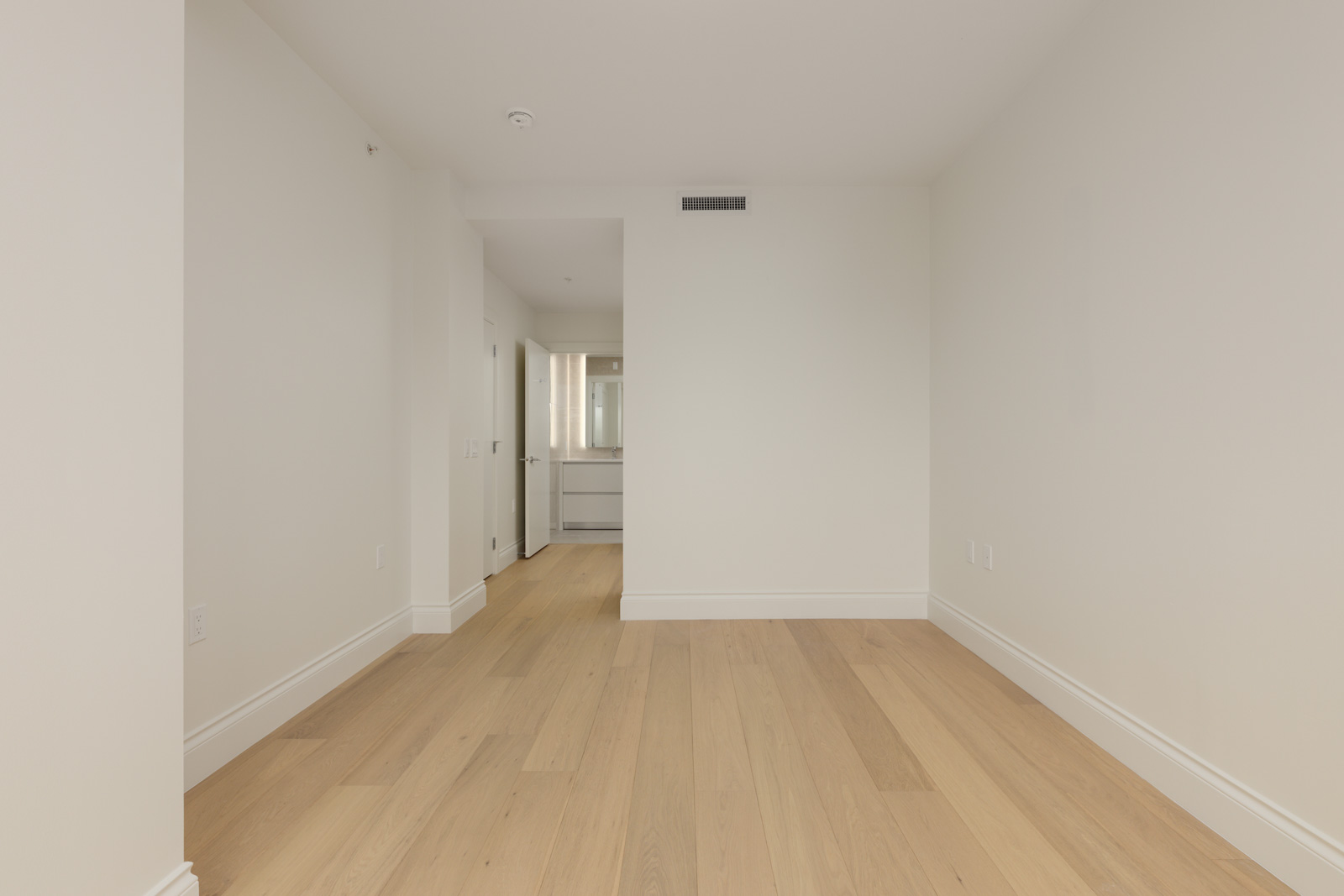 Empty room with light wood flooring, white walls, and an open doorway leading to a bathroom with a large mirror and sink visible in the background.