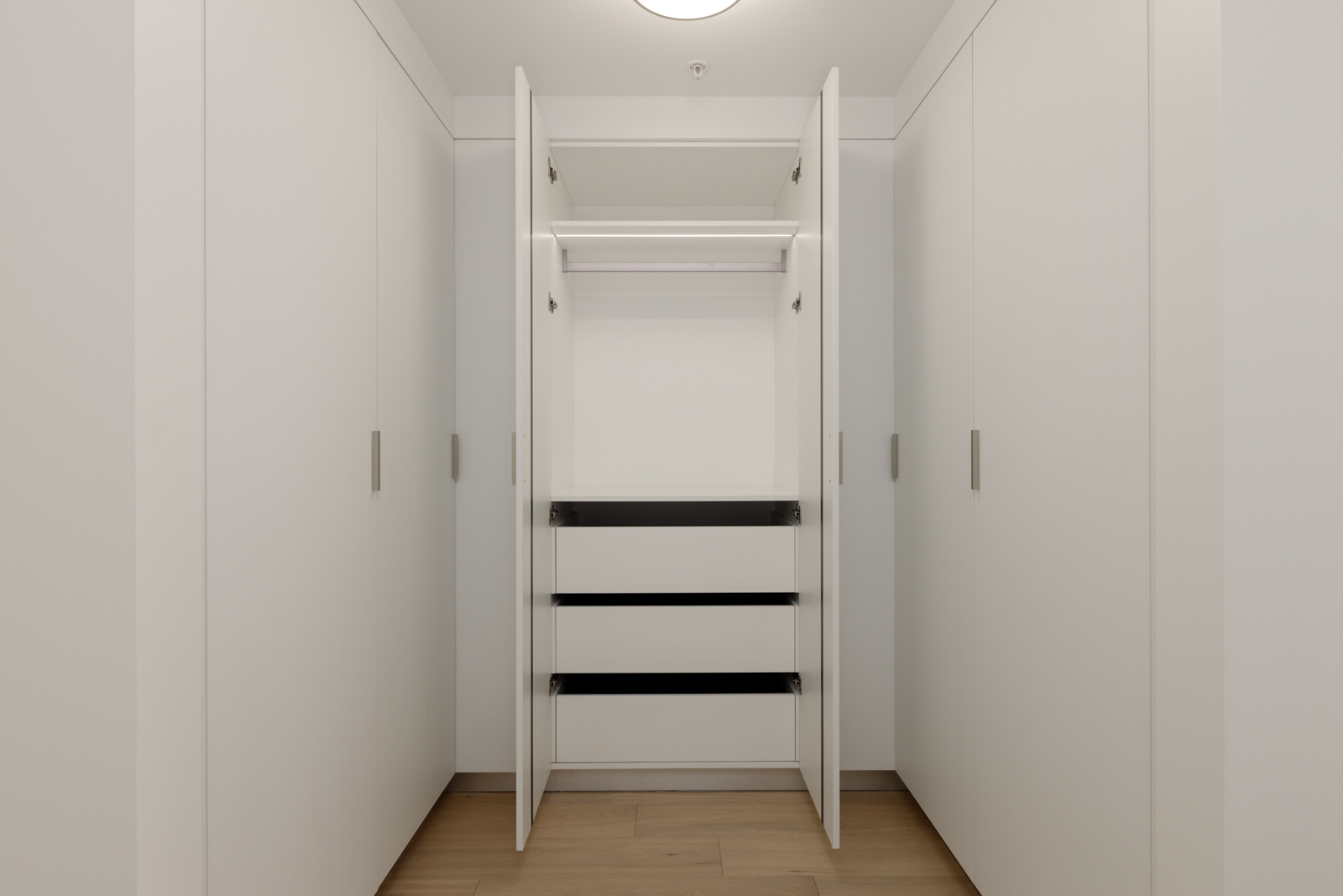 A modern, empty white closet with three open drawers and two open cabinet doors, set against a light wood floor and white walls.