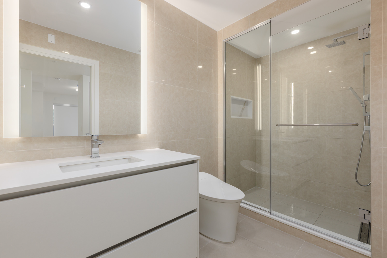 Modern bathroom with a white vanity, built-in sink, illuminated mirror, toilet, and a glass-enclosed walk-in shower with beige tiled walls and floors.